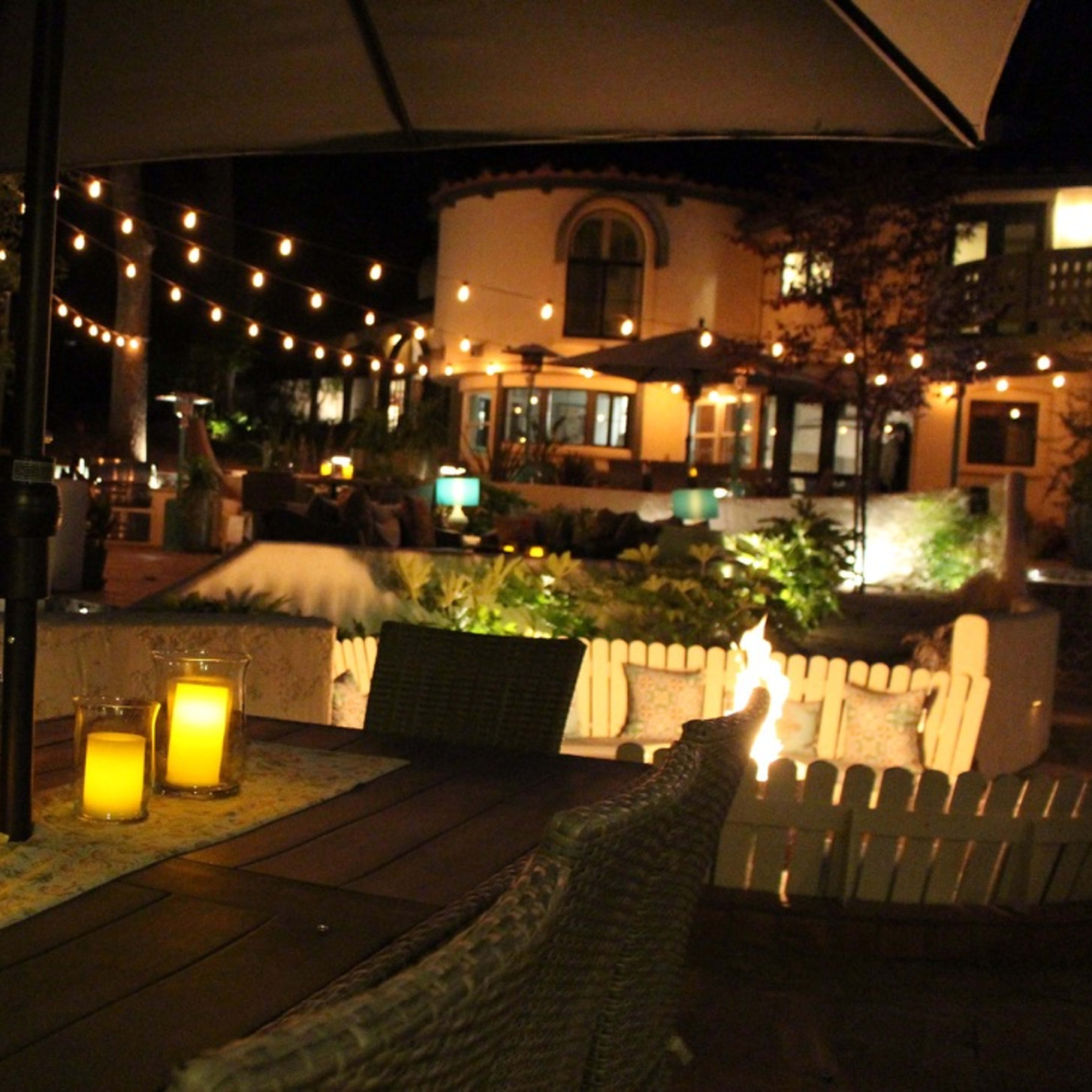 A well-lit outdoor patio features citronella candles on a table, string lights illuminate the surrounding area, and a house is visible in the background.