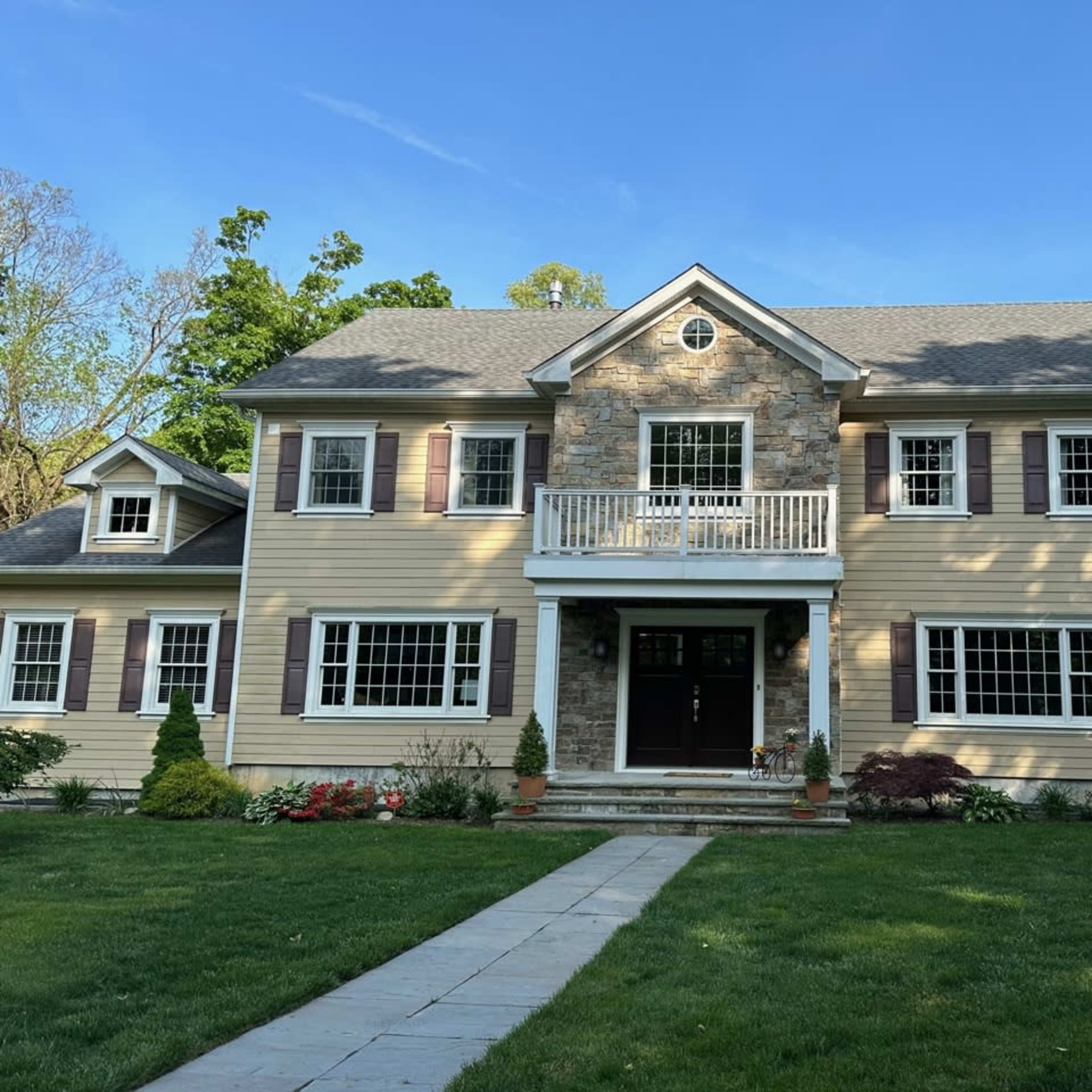 A two-story house with beige siding, stone accents, and a front porch, surrounded by green grass and trees.