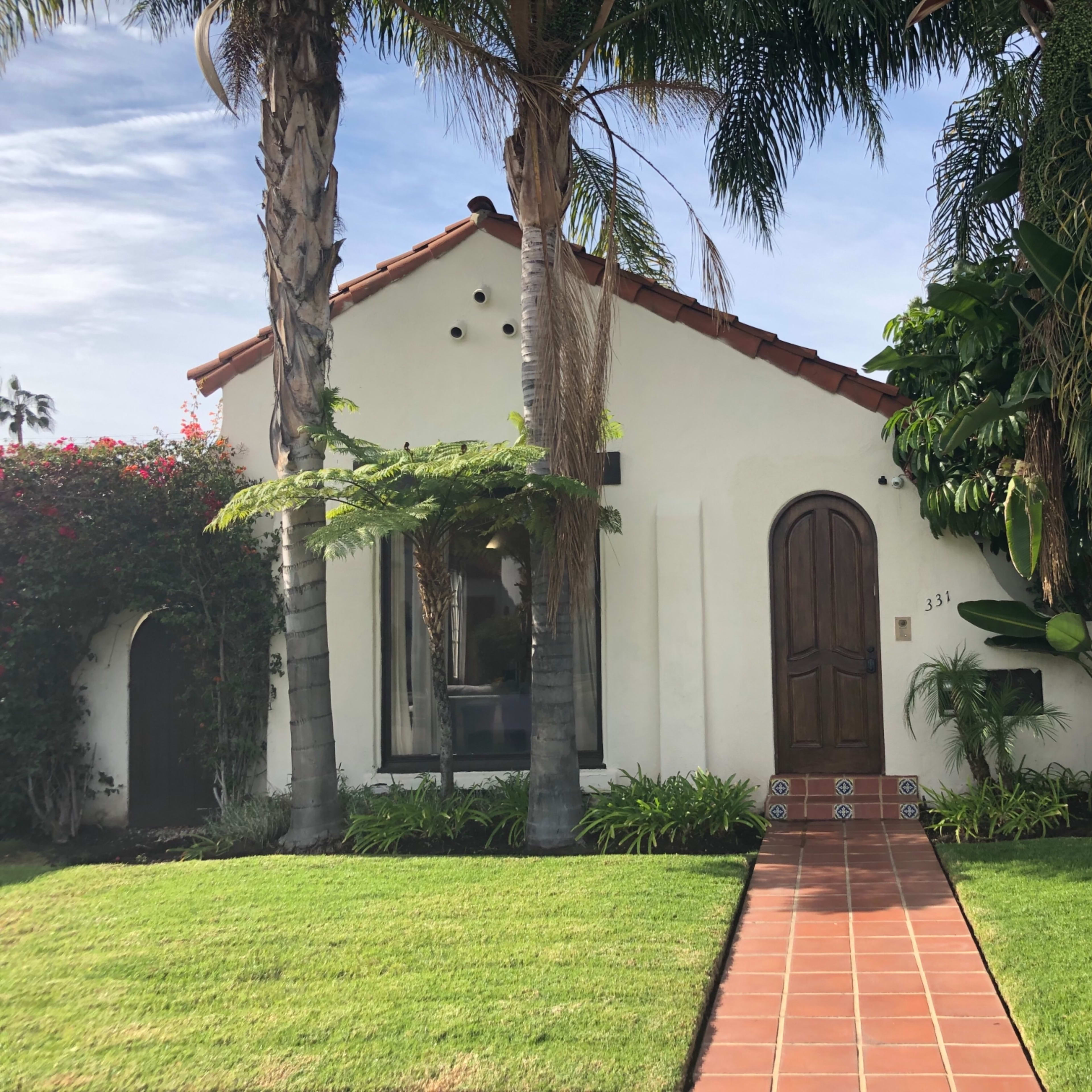 A small, white stucco house with a red-tiled roof is surrounded by palm trees and tropical plants.