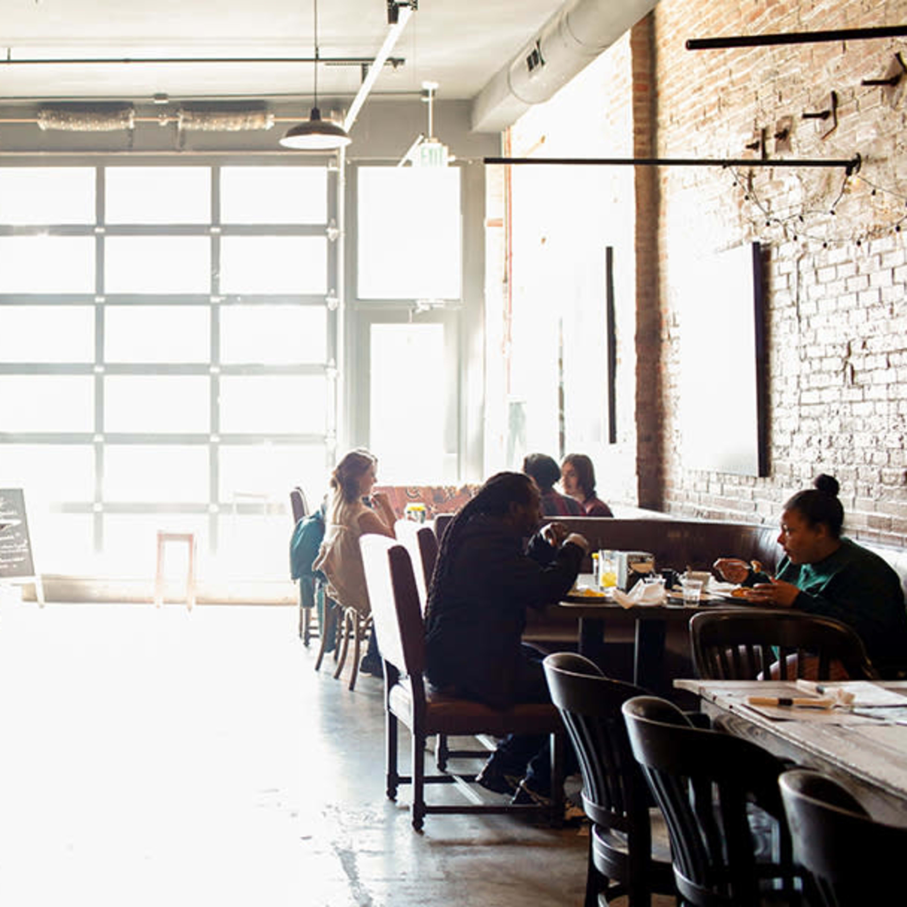 A bright café interior features several tables occupied by patrons, with large windows allowing natural light to flood the space.