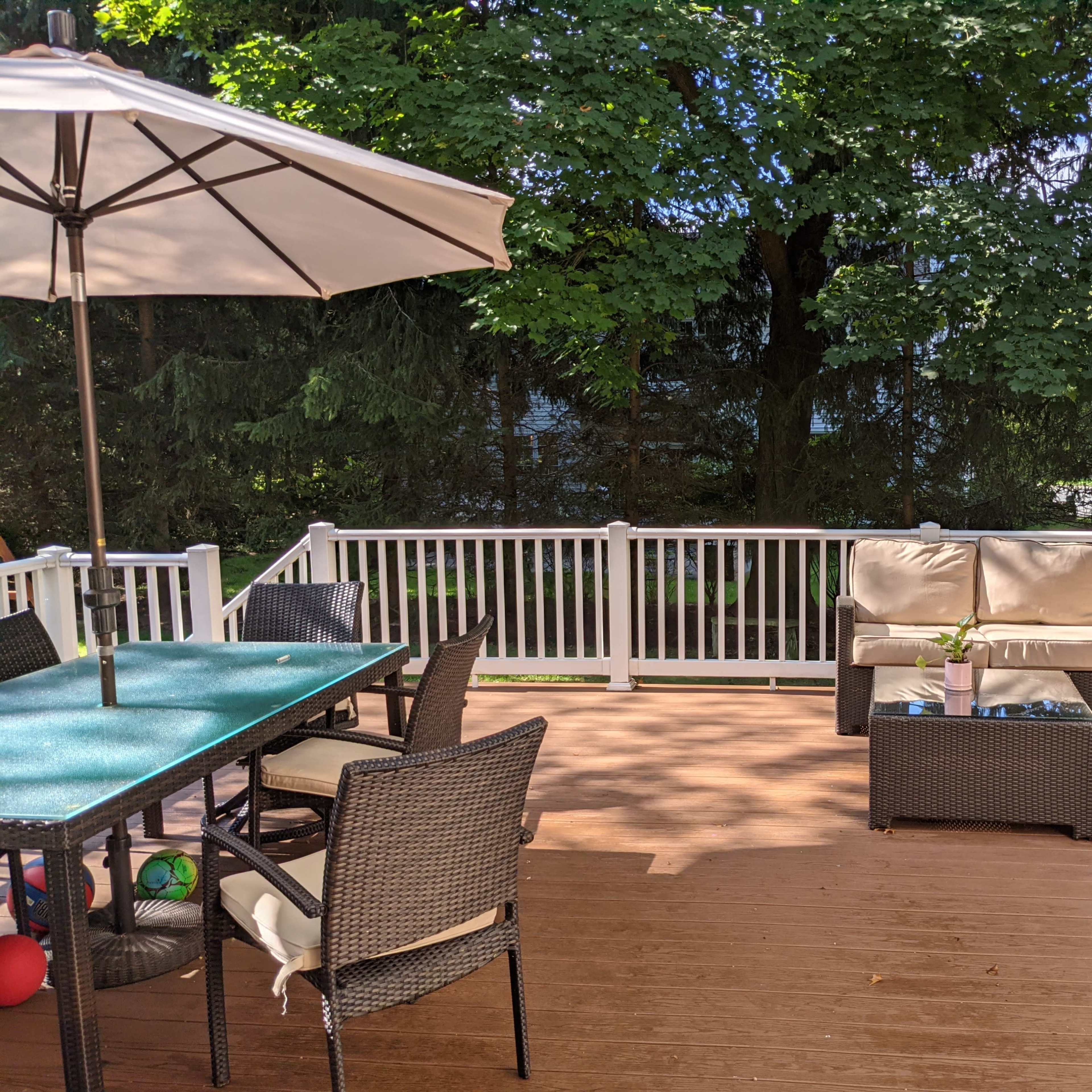 A deck features a table with chairs, a shaded area with an umbrella, and a seating arrangement with cushions in a wooded outdoor setting.