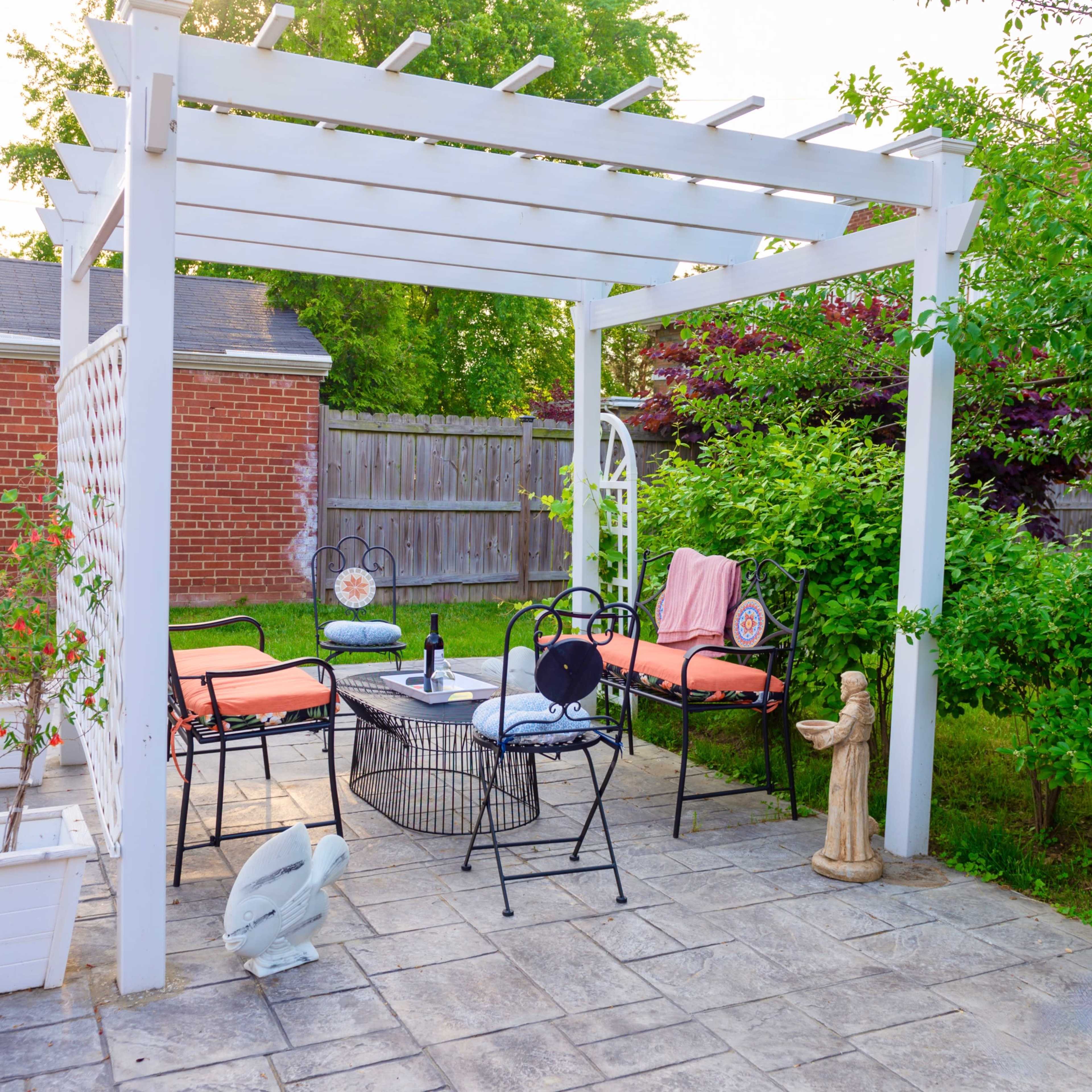 A white pergola with seating and a small table is set up on a stone patio surrounded by greenery.