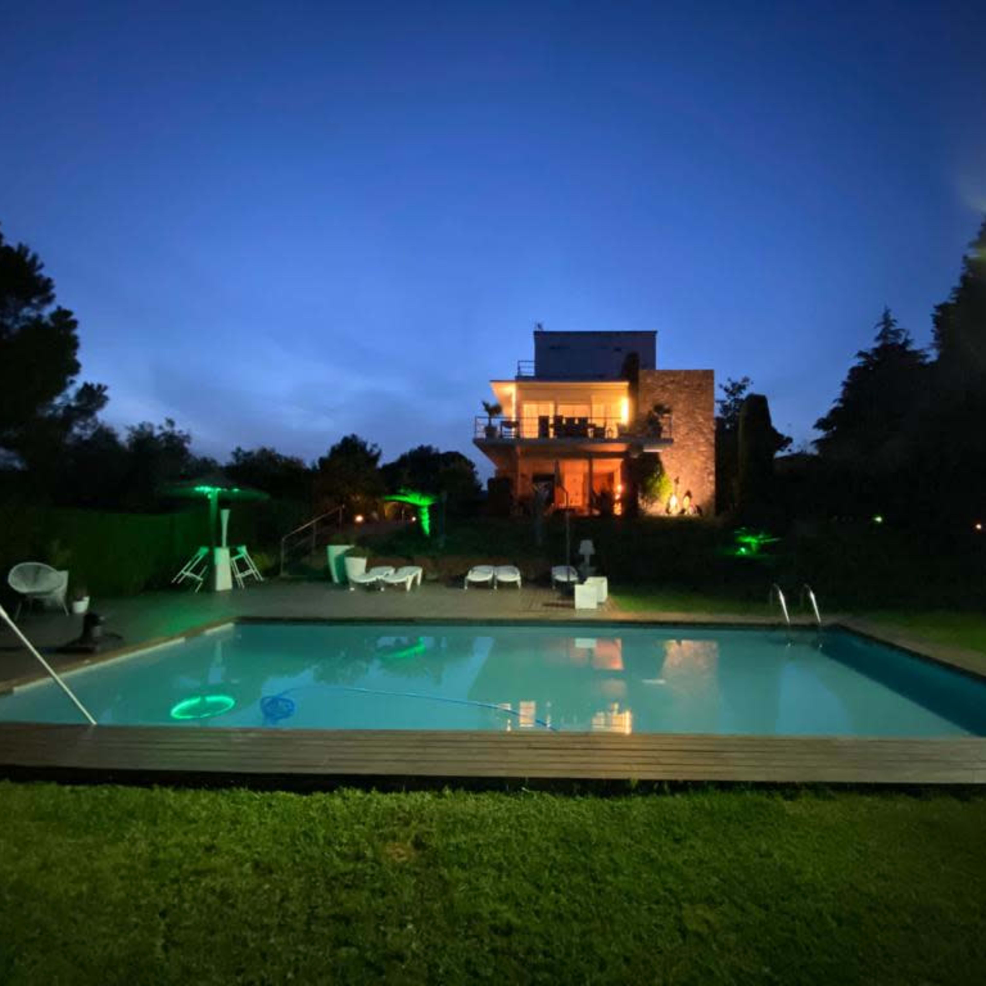 A modern house with stone accents stands behind a well-lit swimming pool surrounded by lush greenery at night.