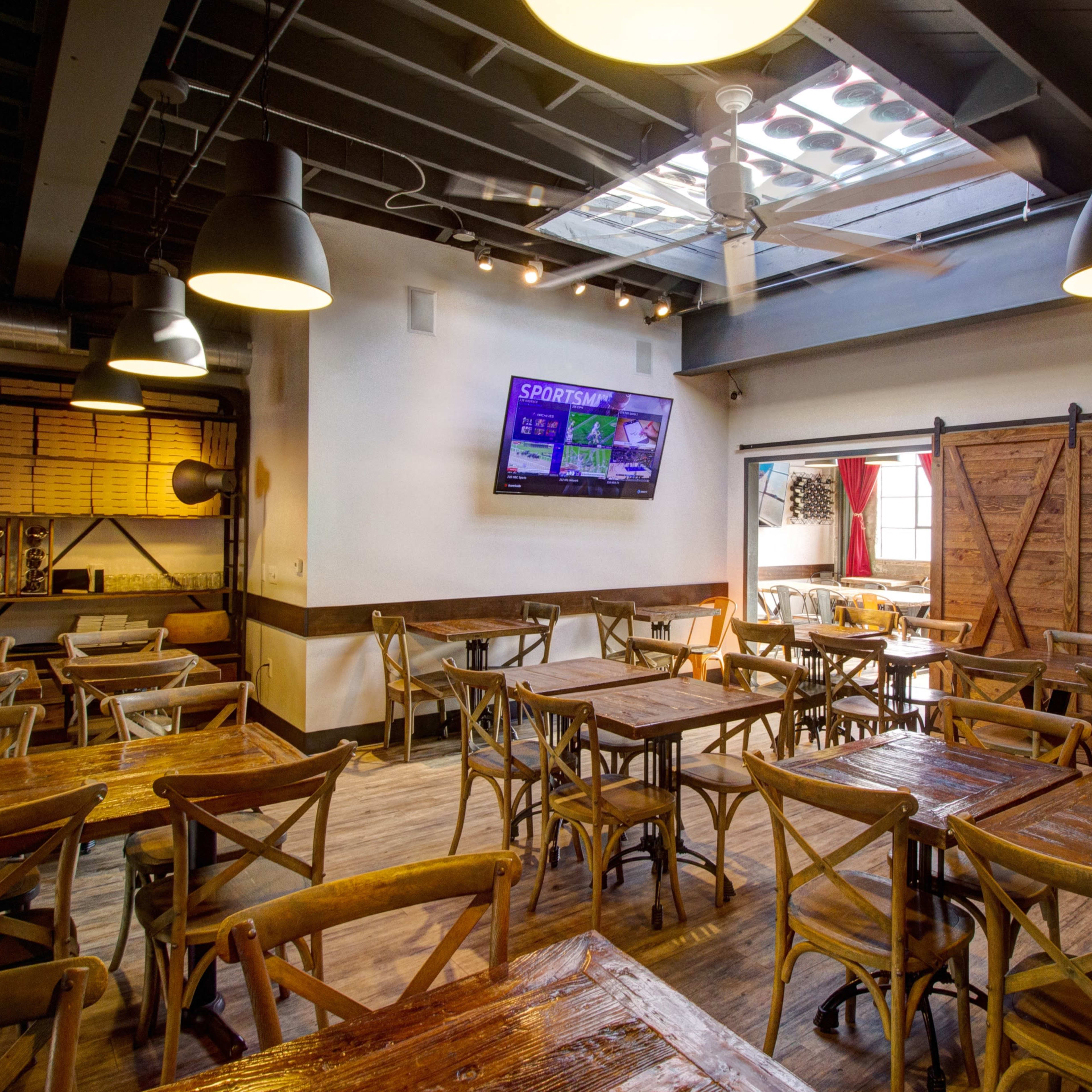 A wooden-furnished restaurant interior with multiple tables, a television on the wall, and a large sliding barn door.