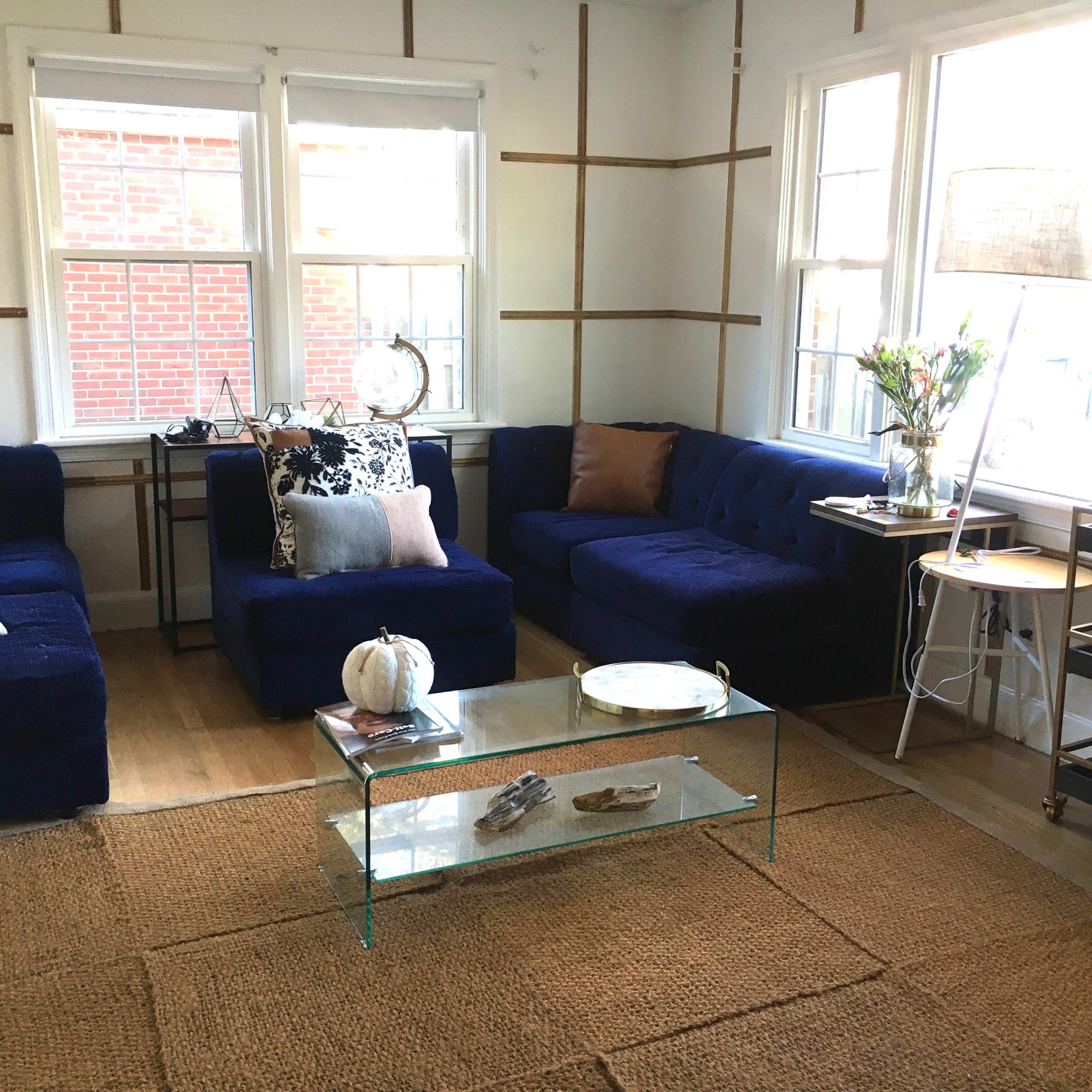 The living room features blue sofas arranged around a glass coffee table, with large windows allowing natural light and a jute rug on the wooden floor.