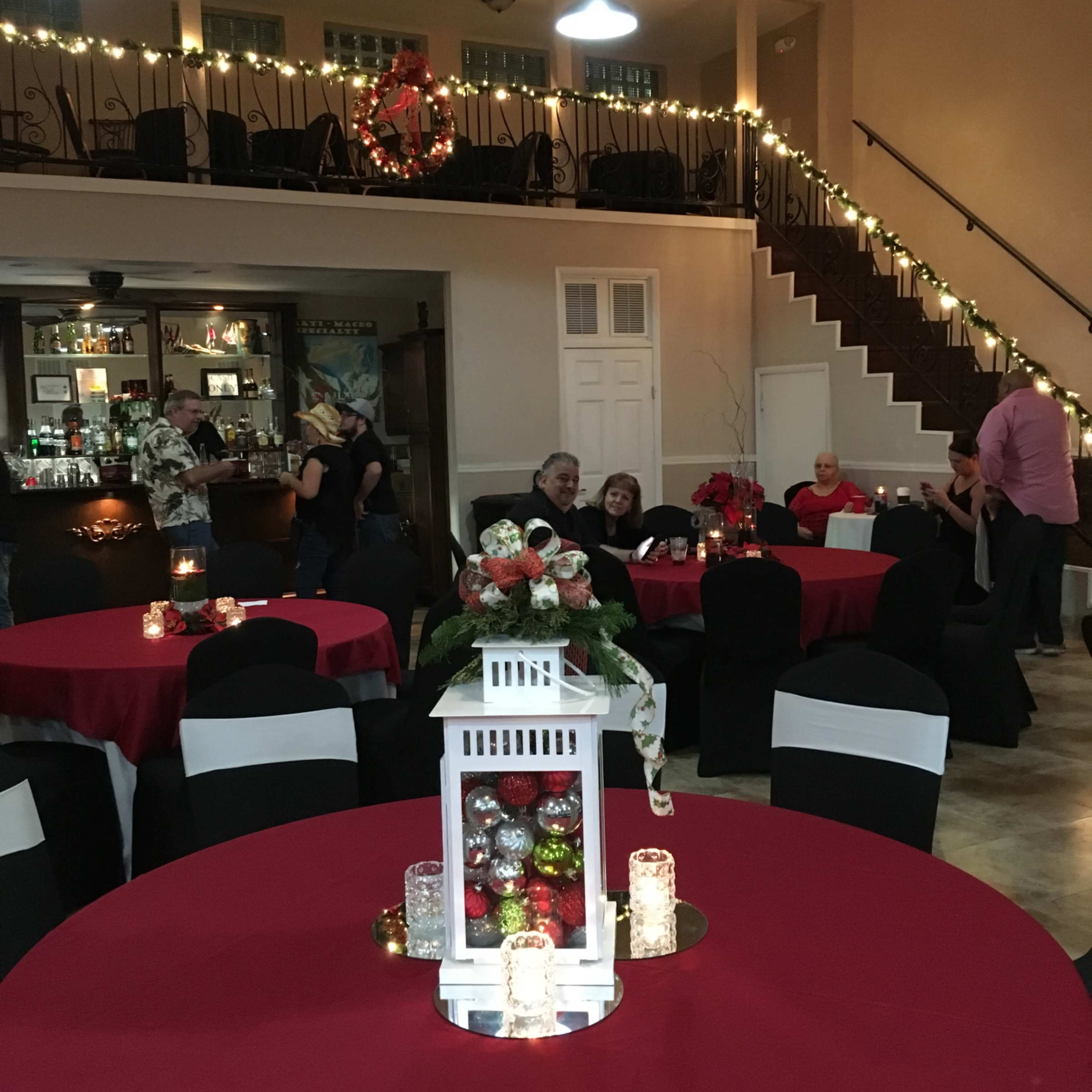 A decorated event space with round tables covered in red tablecloths, black chairs, and festive lighting along the walls and staircase.