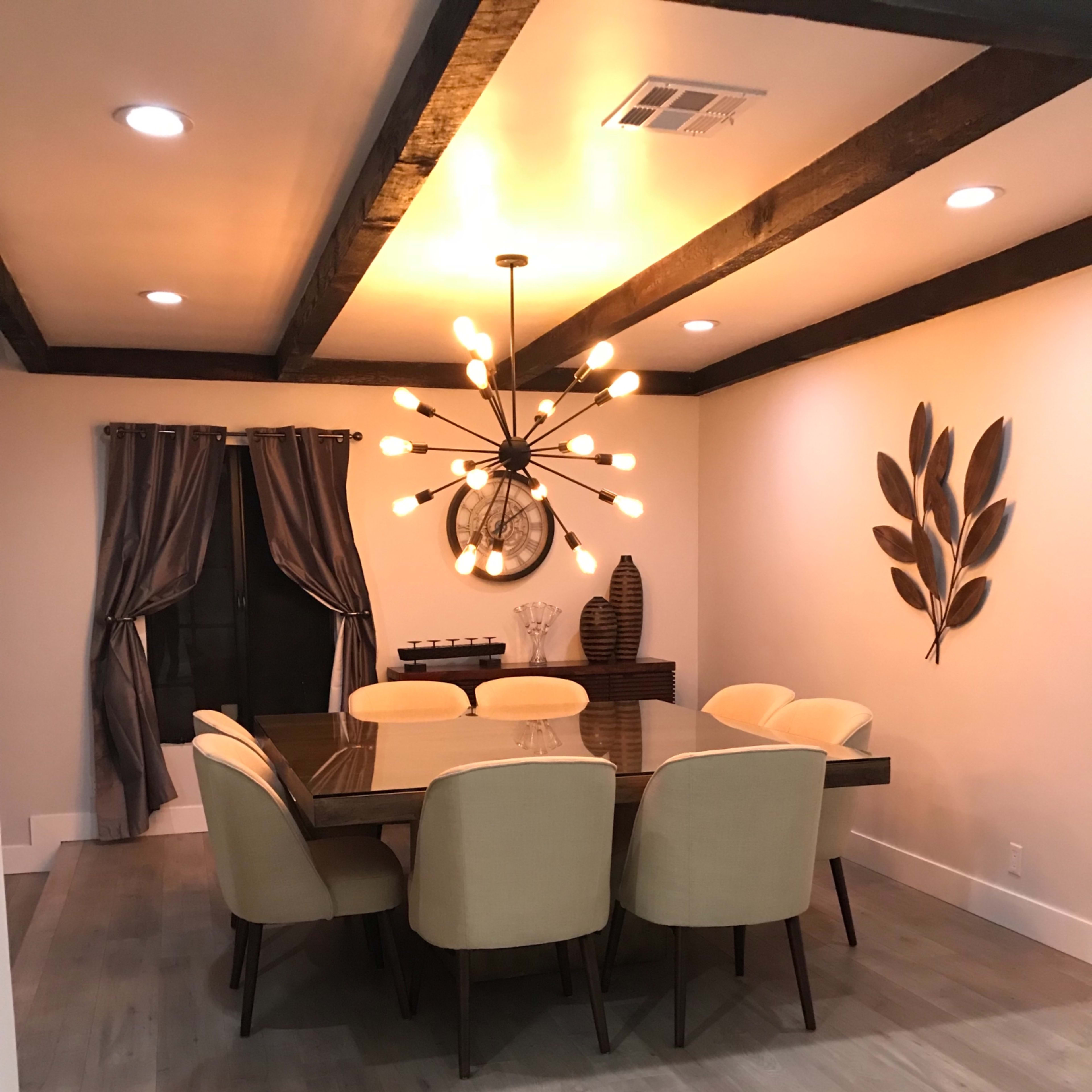 A dining room features a large table surrounded by beige upholstered chairs, with a modern chandelier and decorative wall elements.
