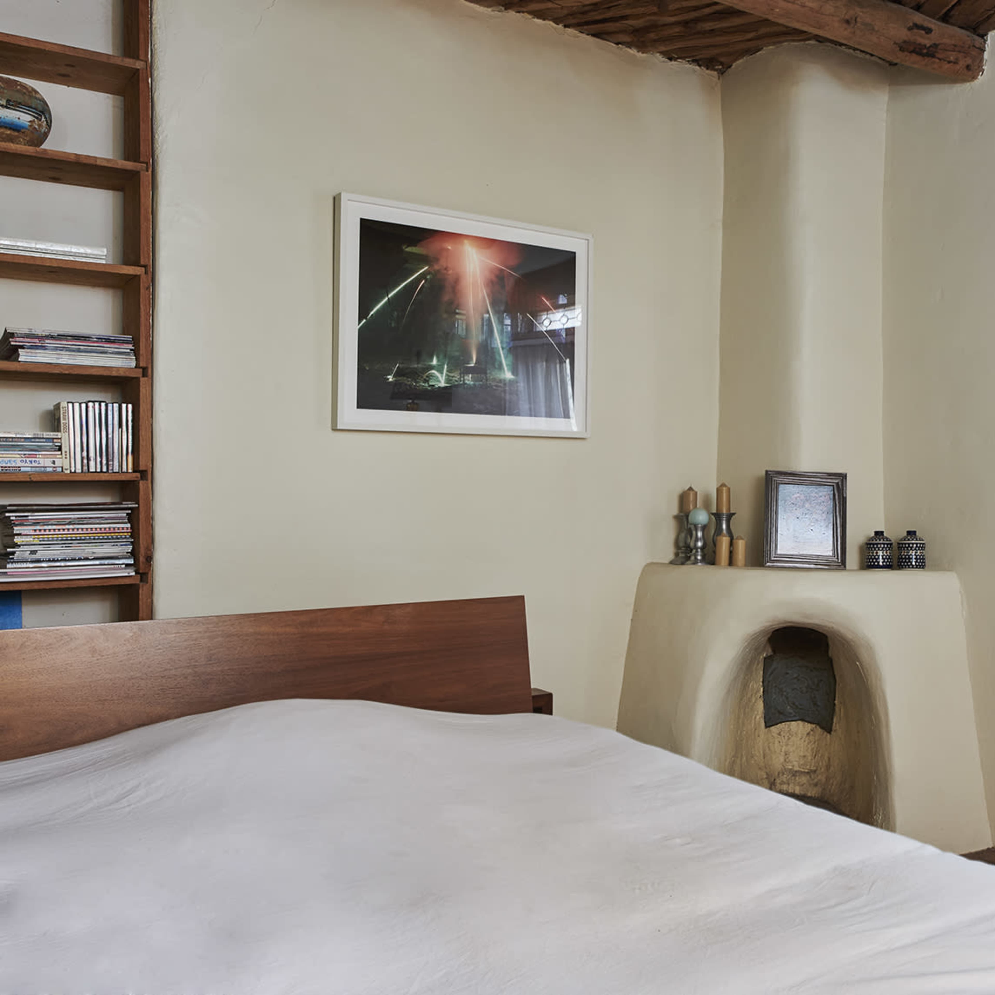 A simple bedroom with a wooden bed, a shelf filled with CDs, and a framed picture on the wall beside a white brick chimney.