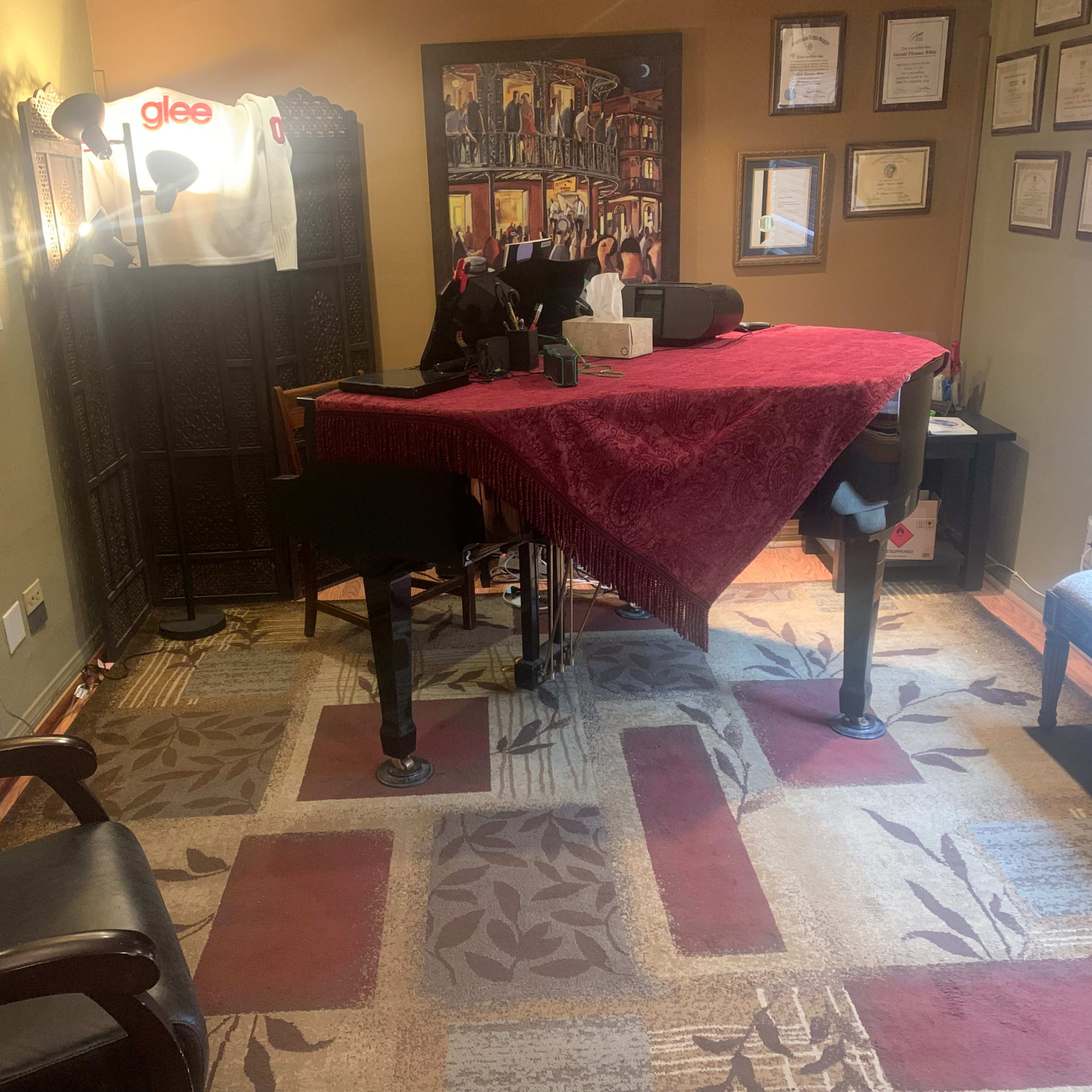 A room with a grand piano covered by a red cloth, a desk with various items, framed certificates on the walls, and a chair in the corner.