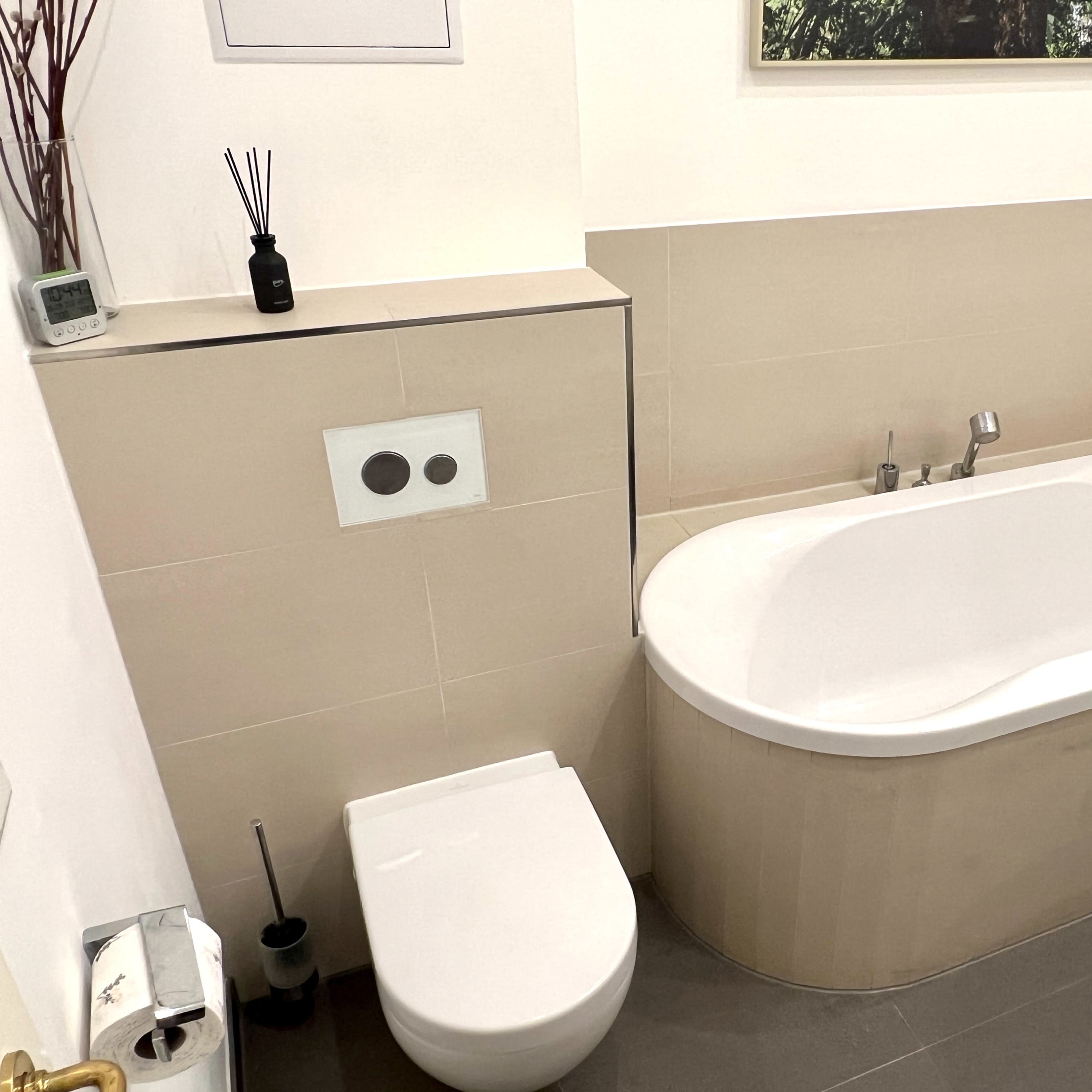A modern bathroom featuring a white freestanding bathtub, a wall-mounted toilet, and neutral-colored tiles.