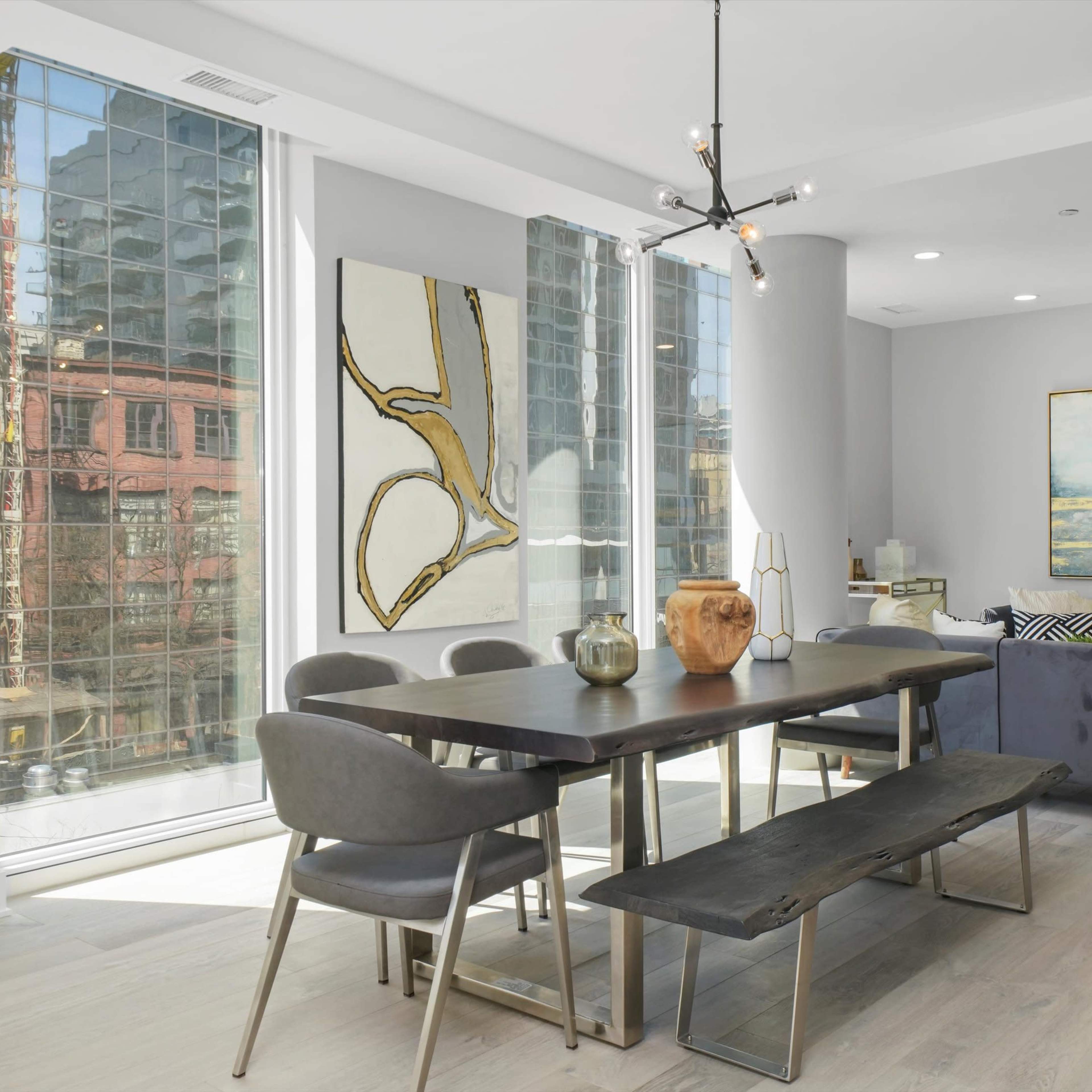A modern dining area with a wooden table, gray chairs, and large windows offering a view of a cityscape.