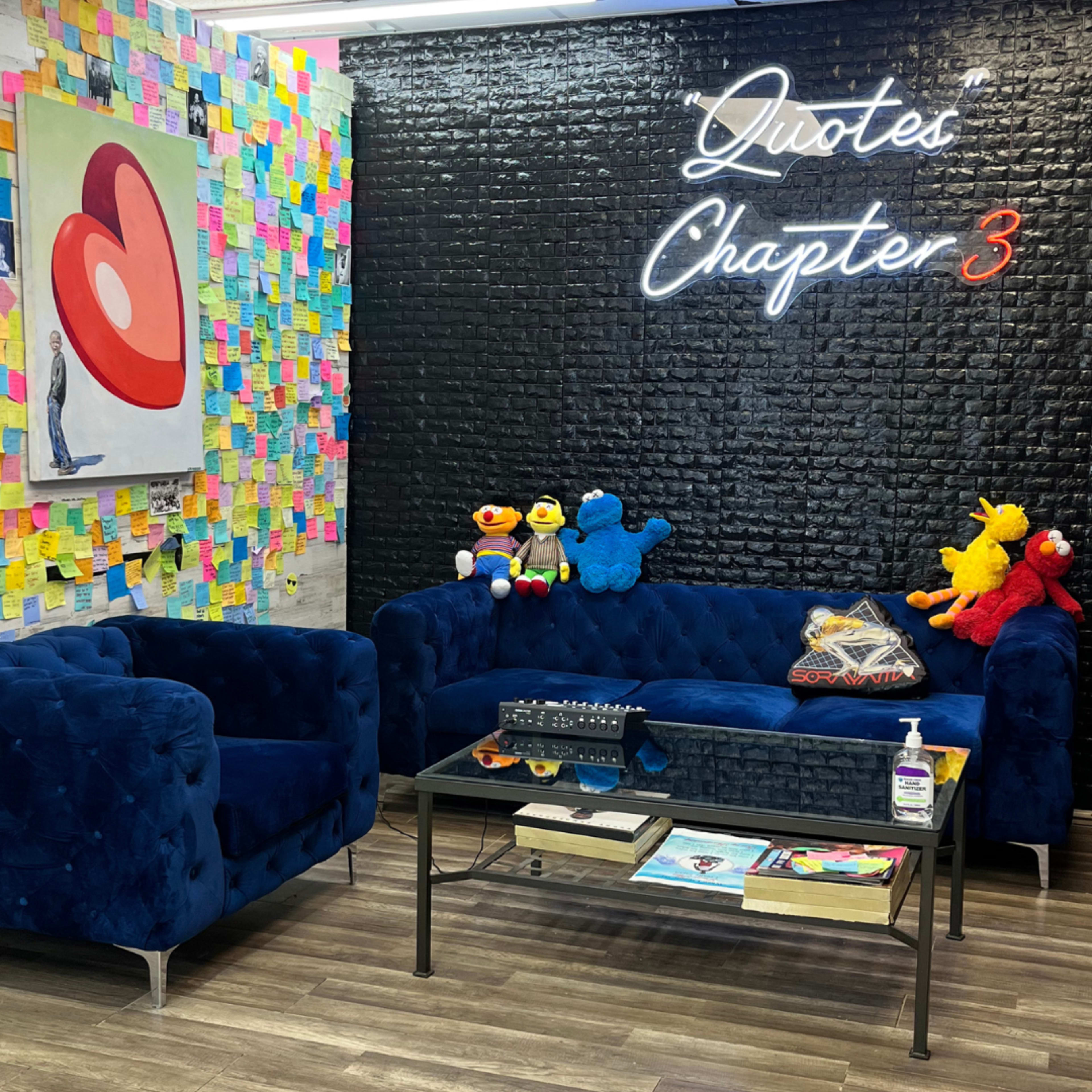 A cozy lounge area with blue sofas, a glass coffee table, colorful sticky notes on the wall, and a neon sign that reads "Quotes Chapter 3."