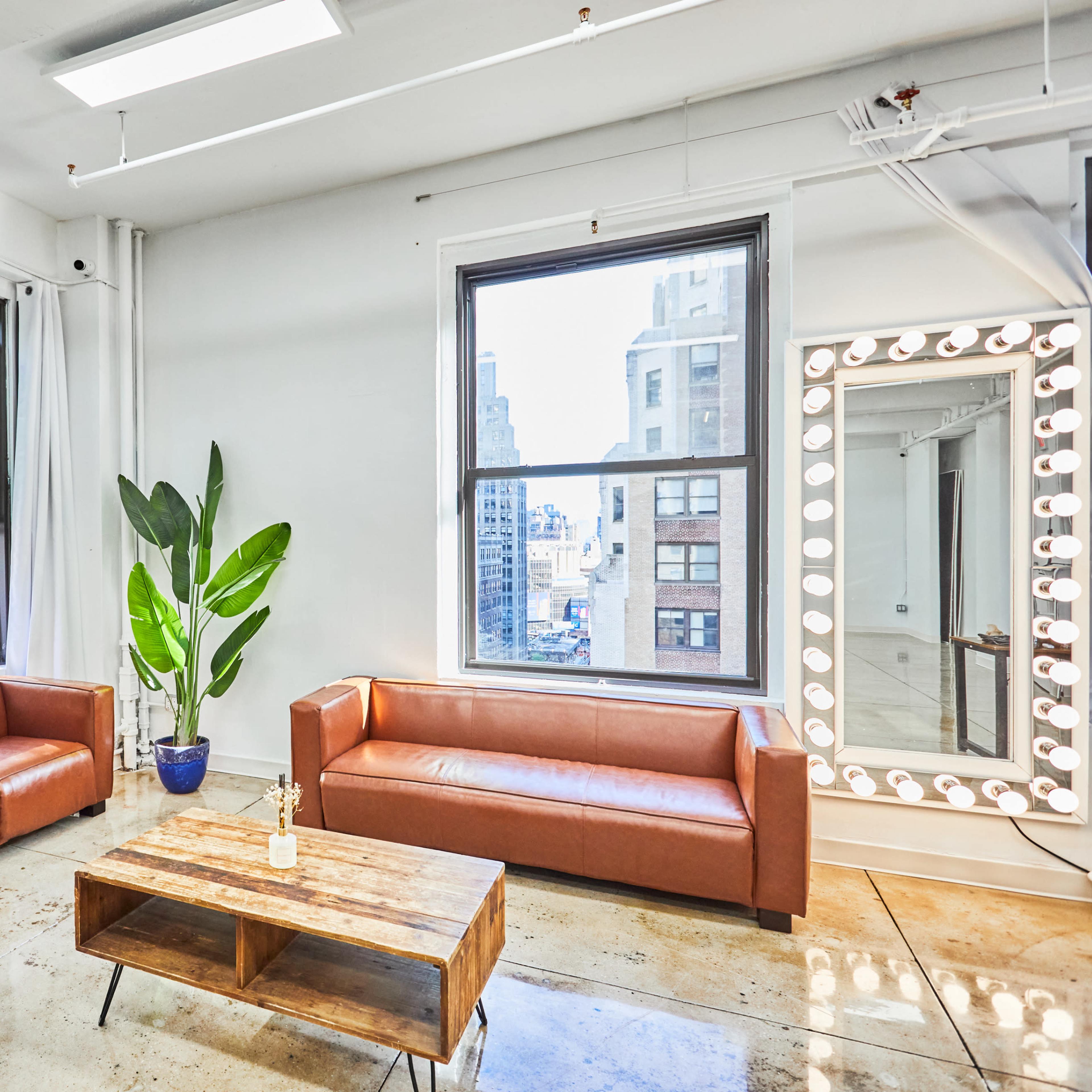 A bright, modern lounge area with two brown leather chairs, a wooden coffee table, and a large mirror with light bulbs, set against large windows overlooking a cityscape.