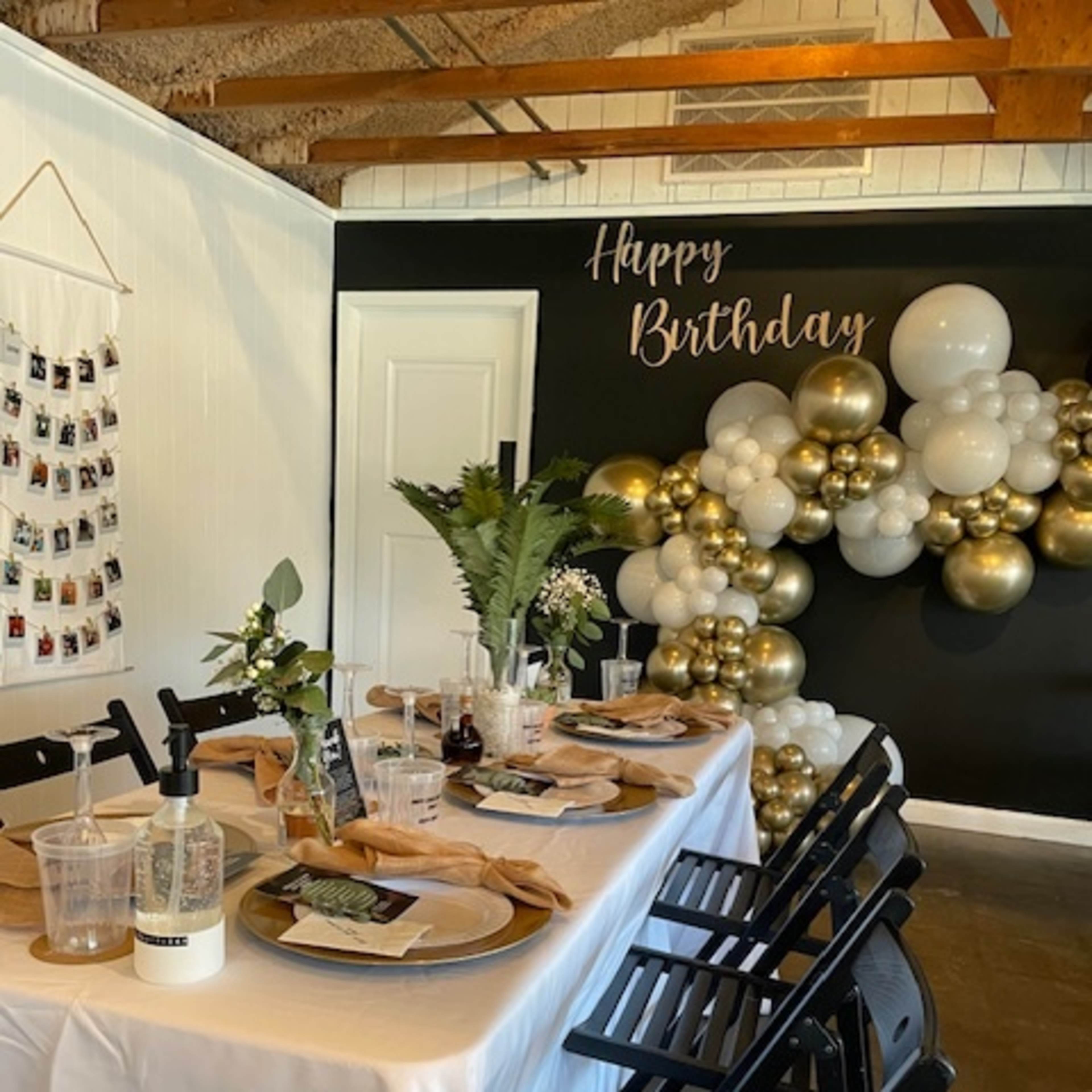 A decorated birthday party table is arranged with plates, glasses, and floral centerpieces against a backdrop of black balloons and a "Happy Birthday" sign.