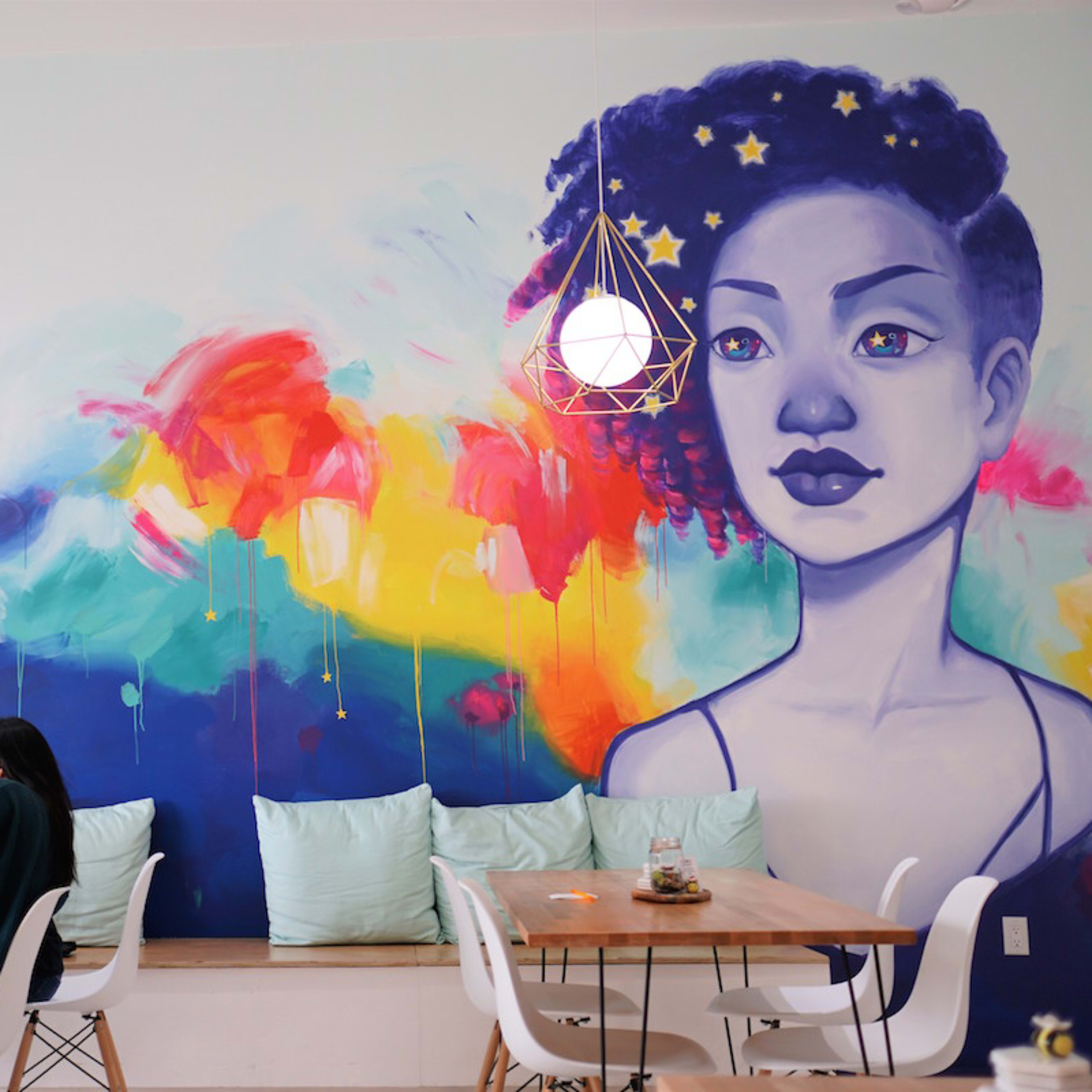 A colorful mural of a woman adorns the wall above a group of three women seated at a wooden table in a cafe.