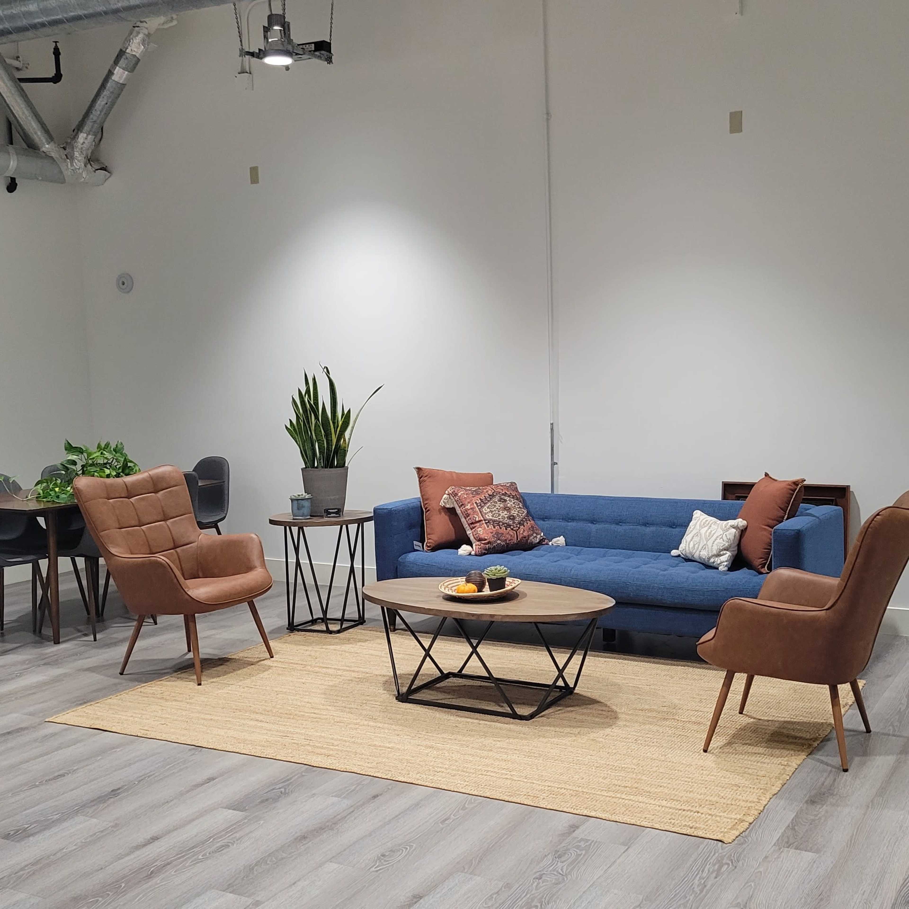 A modern living room features a blue couch, two brown armchairs, a round coffee table with fruit, a decorative rug, and a dining area with a plant.