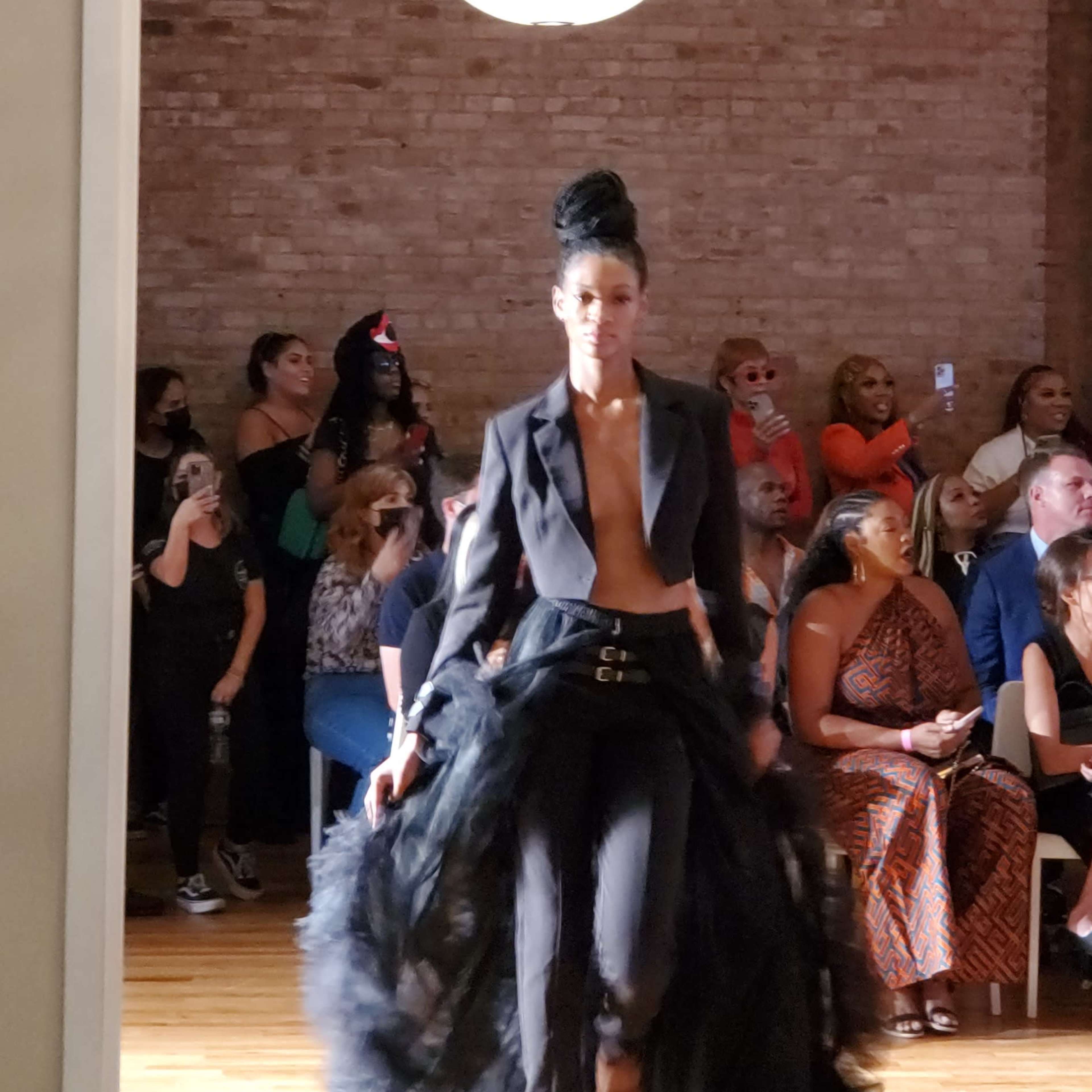 A model walks down the runway wearing a black outfit with a flowing skirt, while an audience watches from the sides.