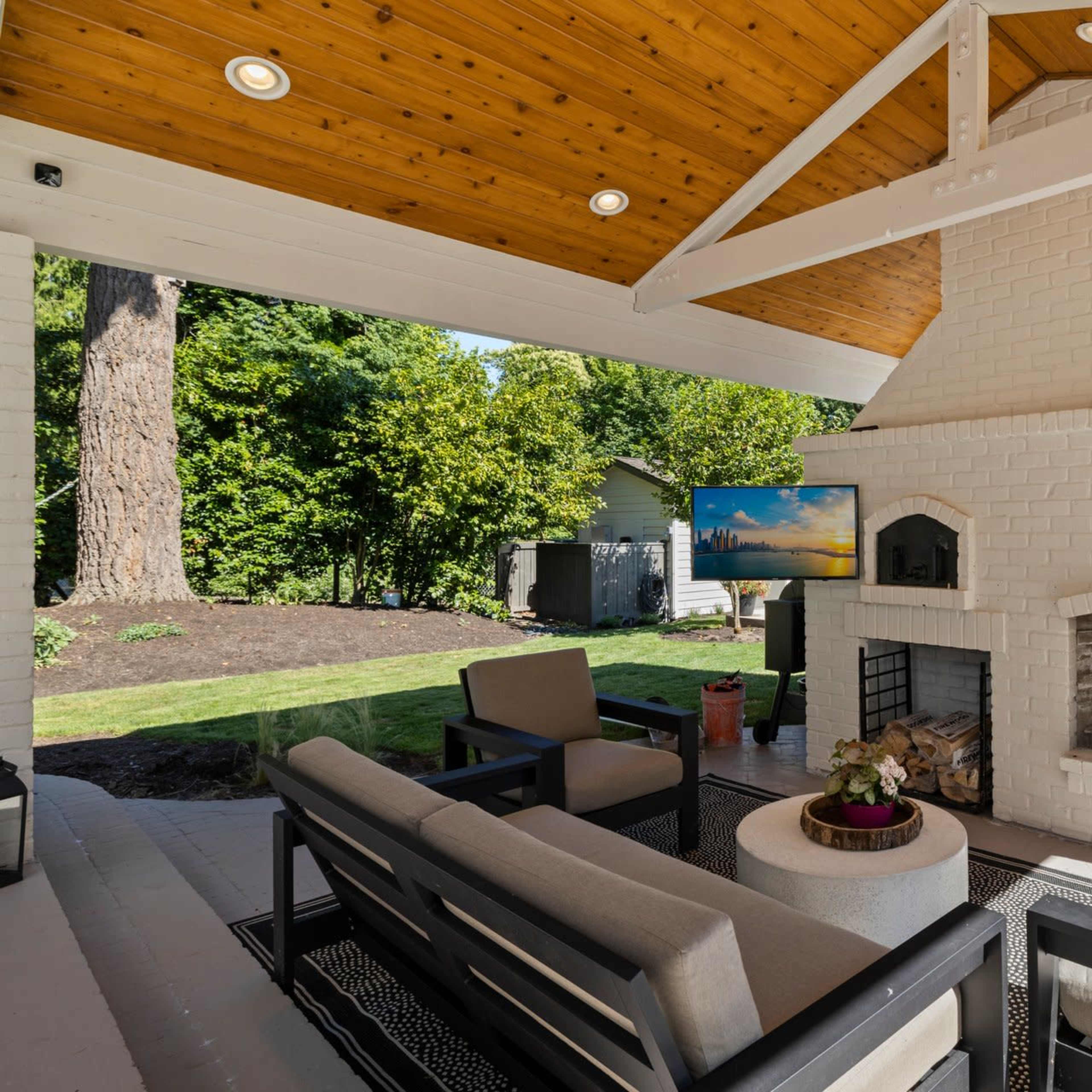 The scene features a covered outdoor patio with a white brick fireplace, modern seating arrangements, and a television mounted on the wall.