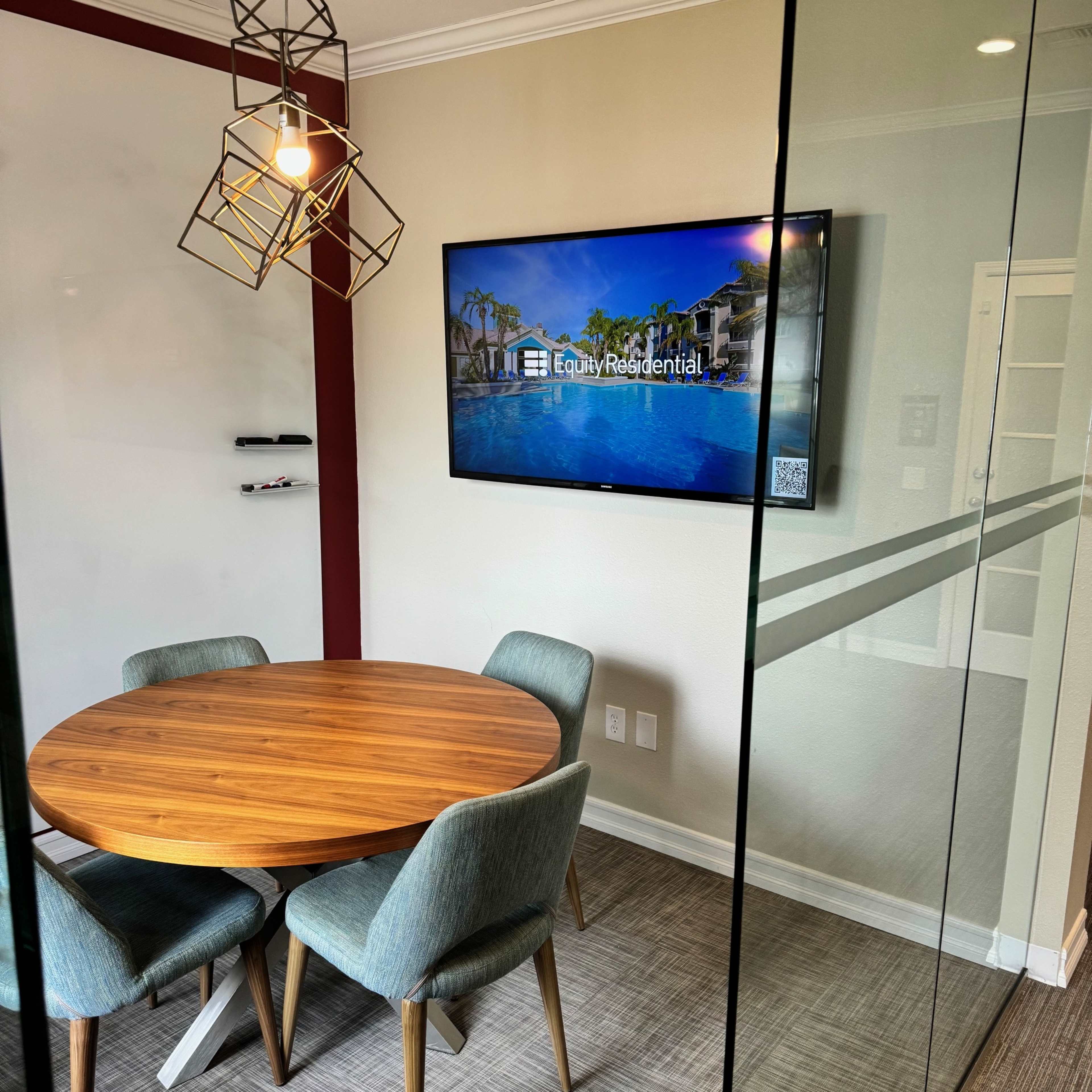 A modern conference room with a round wooden table, four upholstered chairs, and a wall-mounted television displaying a property listing.