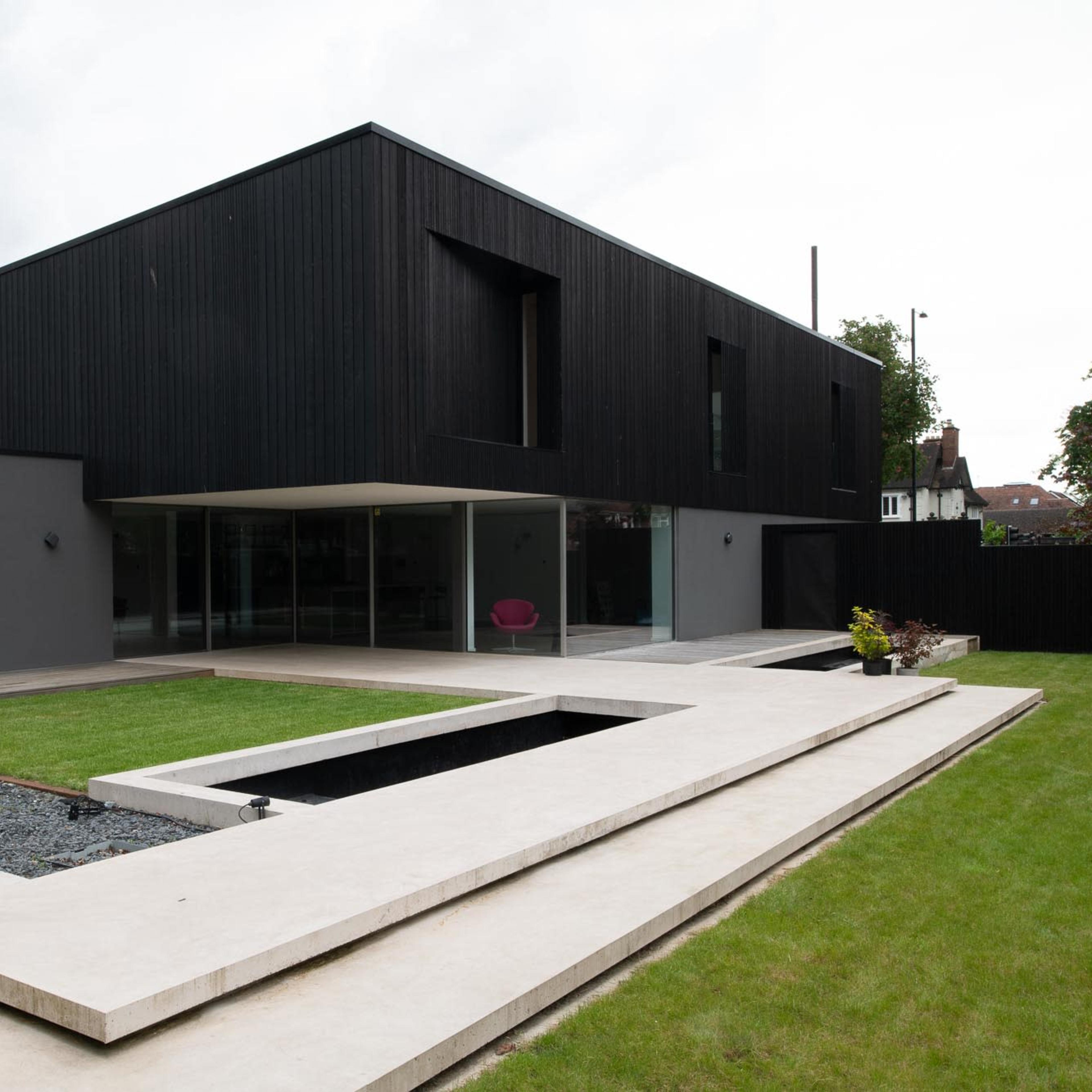 A modern black wooden house with large glass windows is surrounded by a landscaped yard featuring a rectangular stone path and a small water feature.