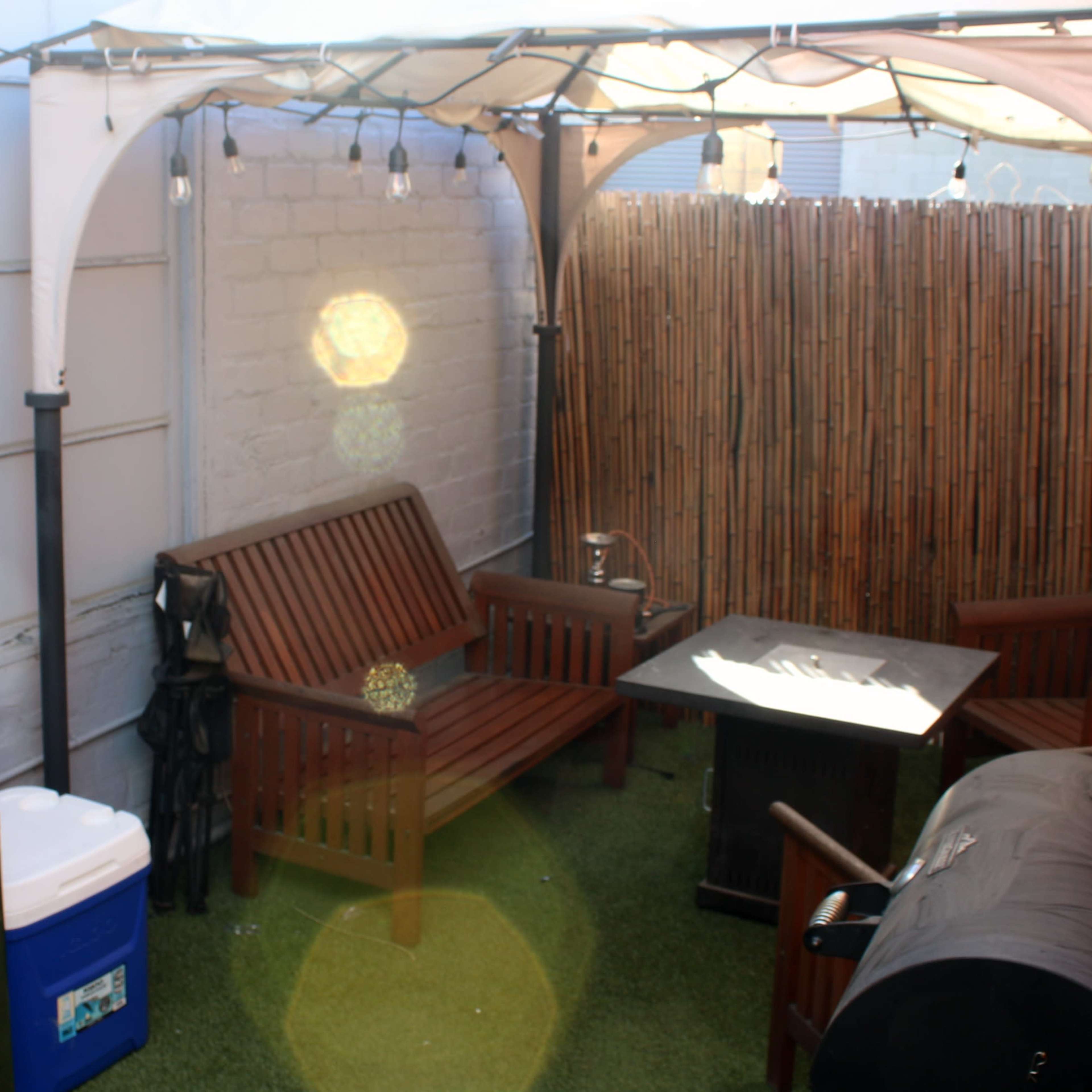 A small outdoor patio area with a wooden bench, a table, a charcoal grill, and a cooler, all under a shaded canopy.