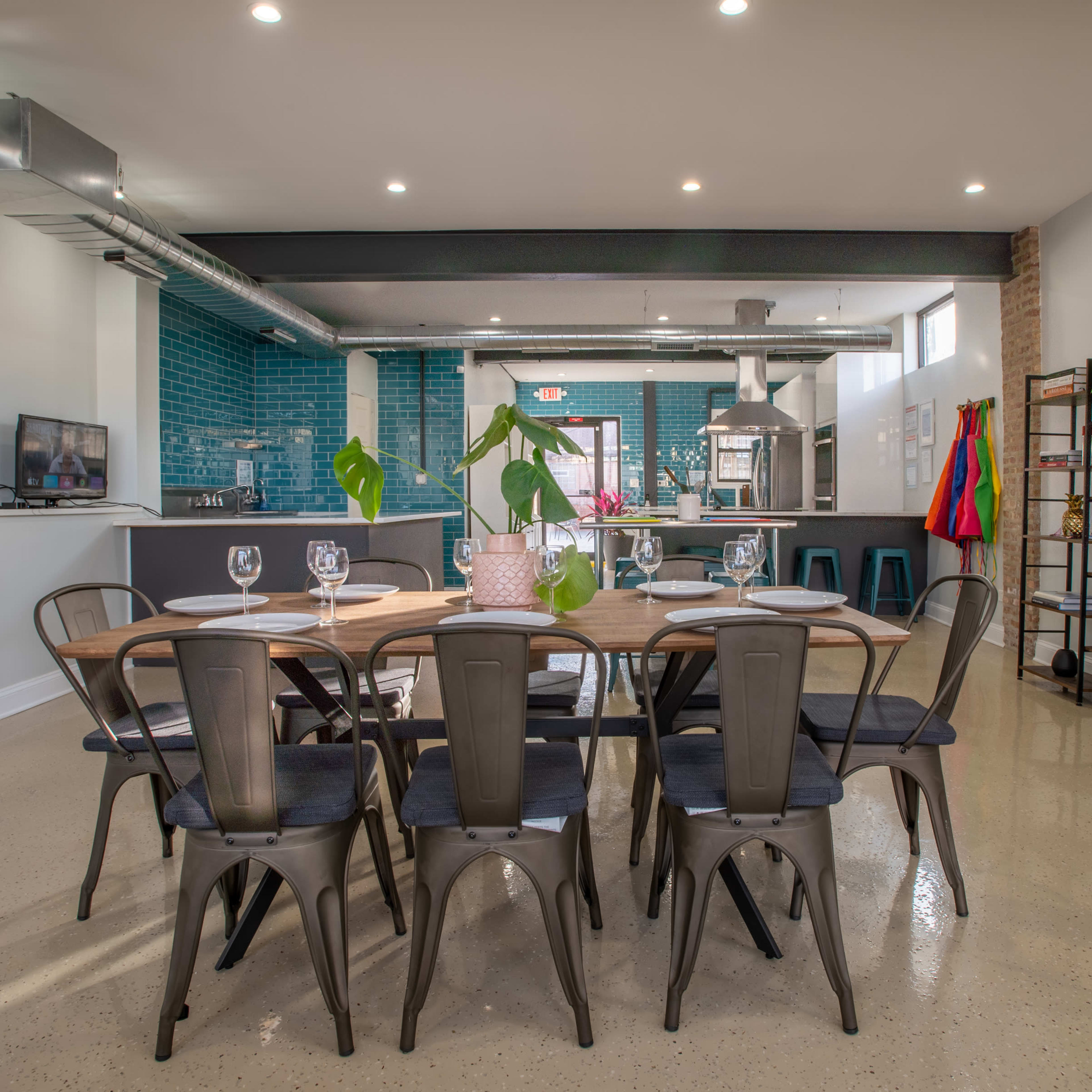 A modern dining area with a large wooden table surrounded by metal chairs, set against a backdrop of teal tiles and a kitchen workspace.