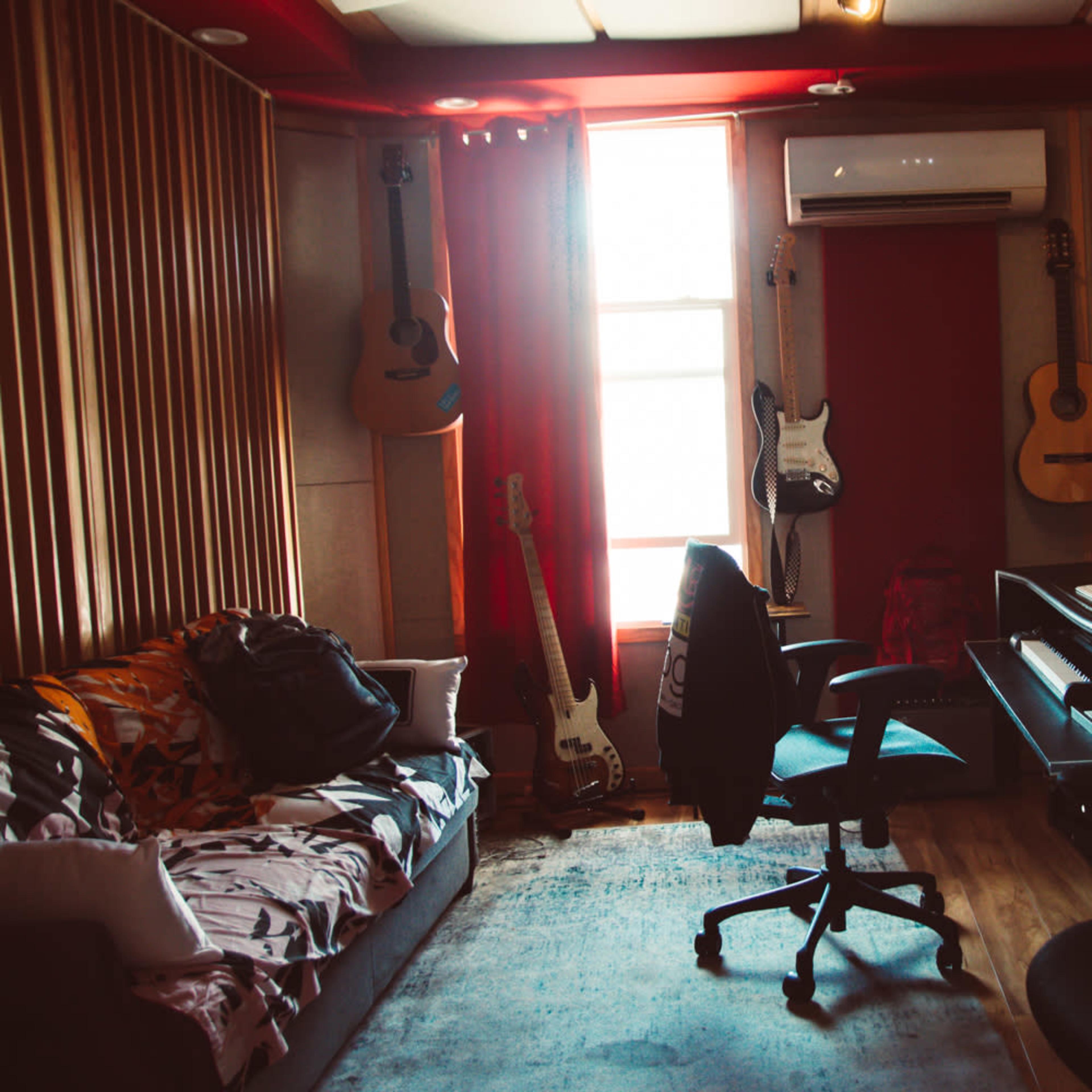 A music studio features a couch, various guitars on the walls, a piano, and recording equipment, with large windows providing natural light.