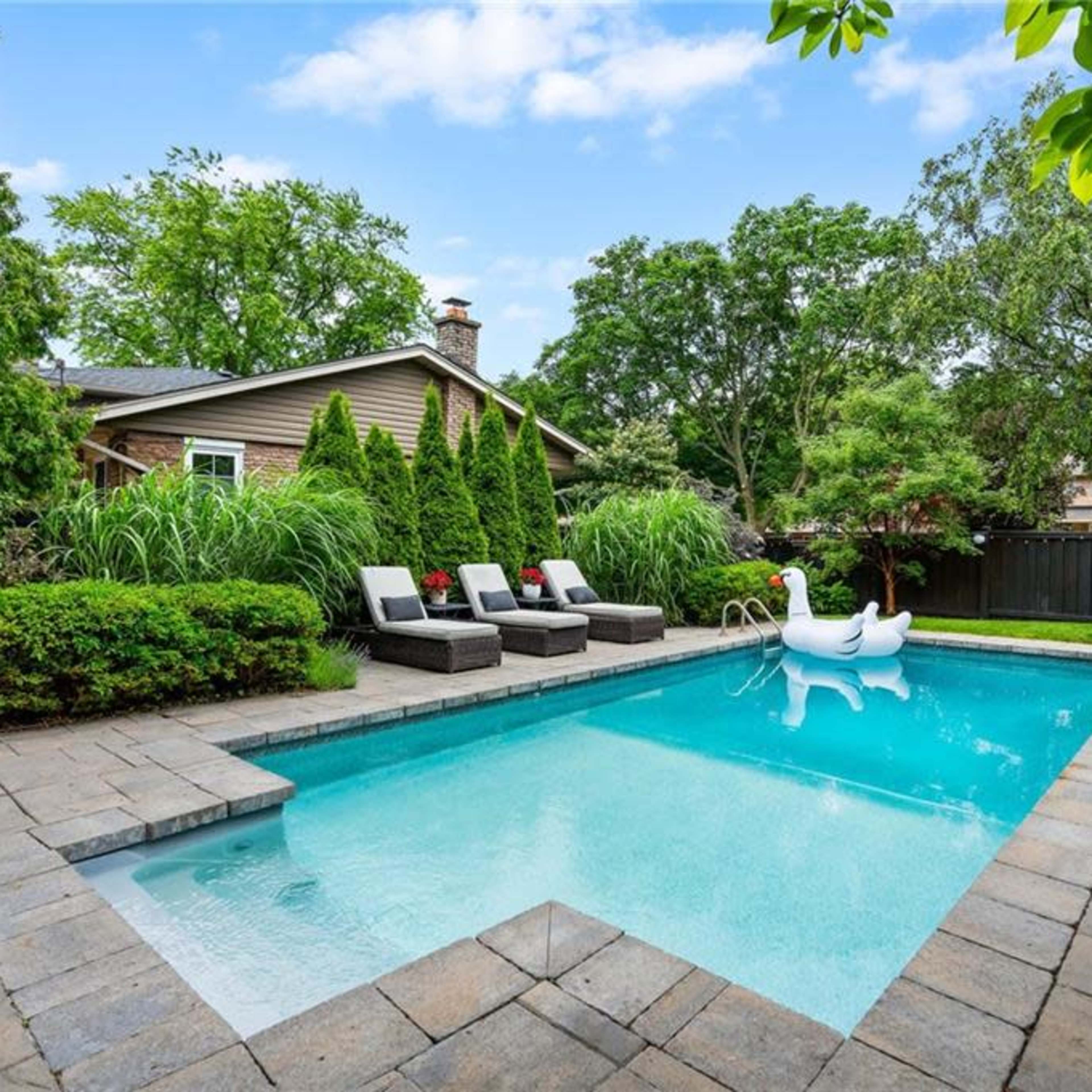 A landscaped backyard with a rectangular swimming pool, surrounded by stone patio, lounge chairs, and decorative greenery.