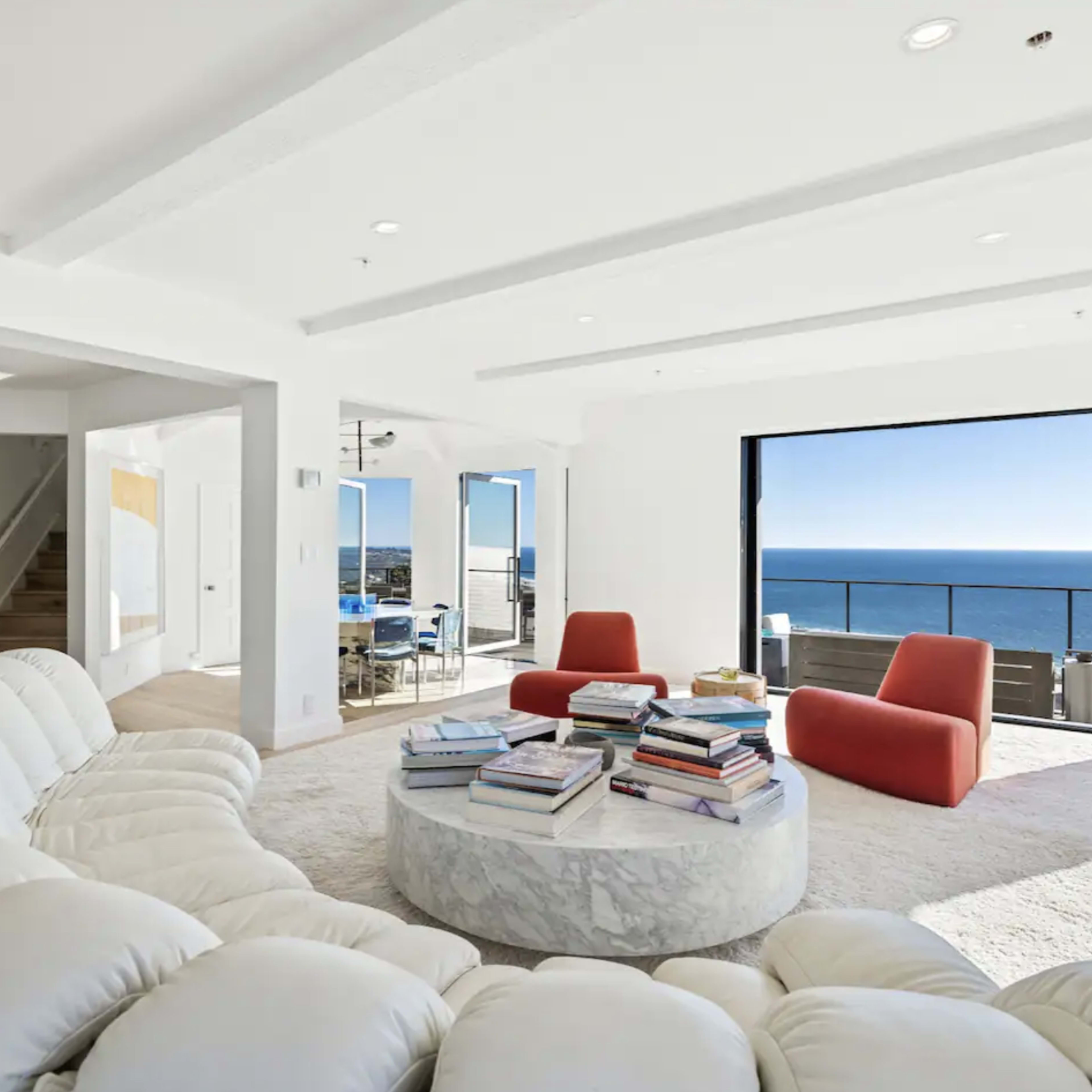 A modern living room features a circular sofa, a marble coffee table stacked with books, large windows overlooking the ocean, and two accent chairs.