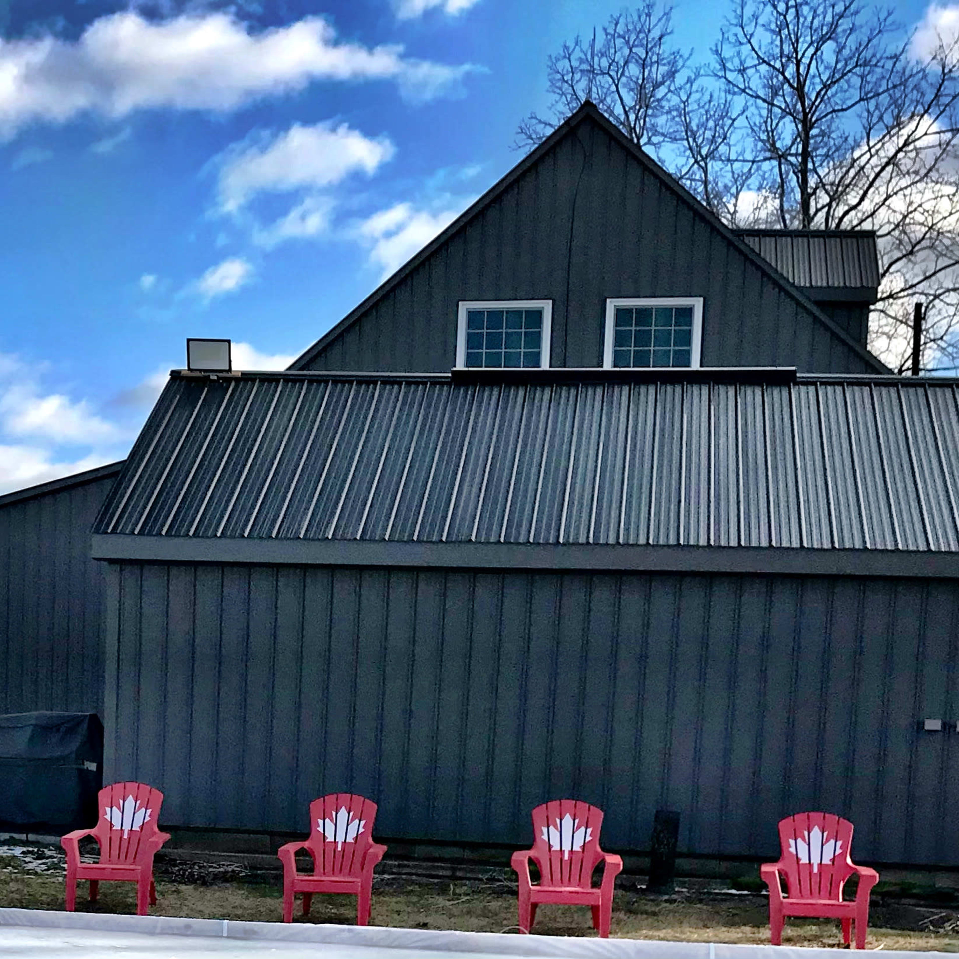 A row of red Adirondack chairs is positioned in front of a gray barn-style building under a partly cloudy sky.