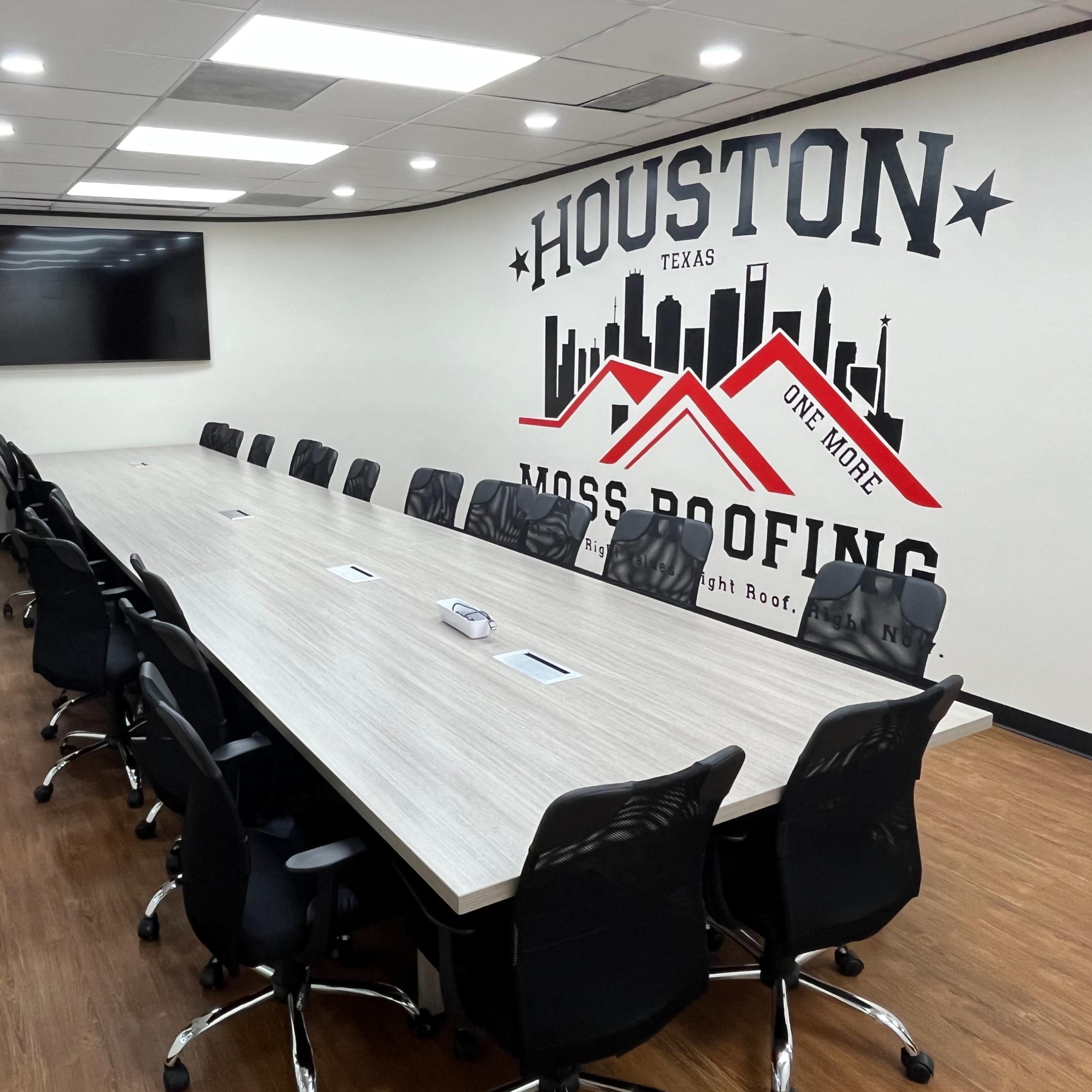A large conference room with a long table surrounded by black chairs, featuring a wall with a large mural that includes the words "HOUSTON TEXAS" and "MOSS ROOFING."