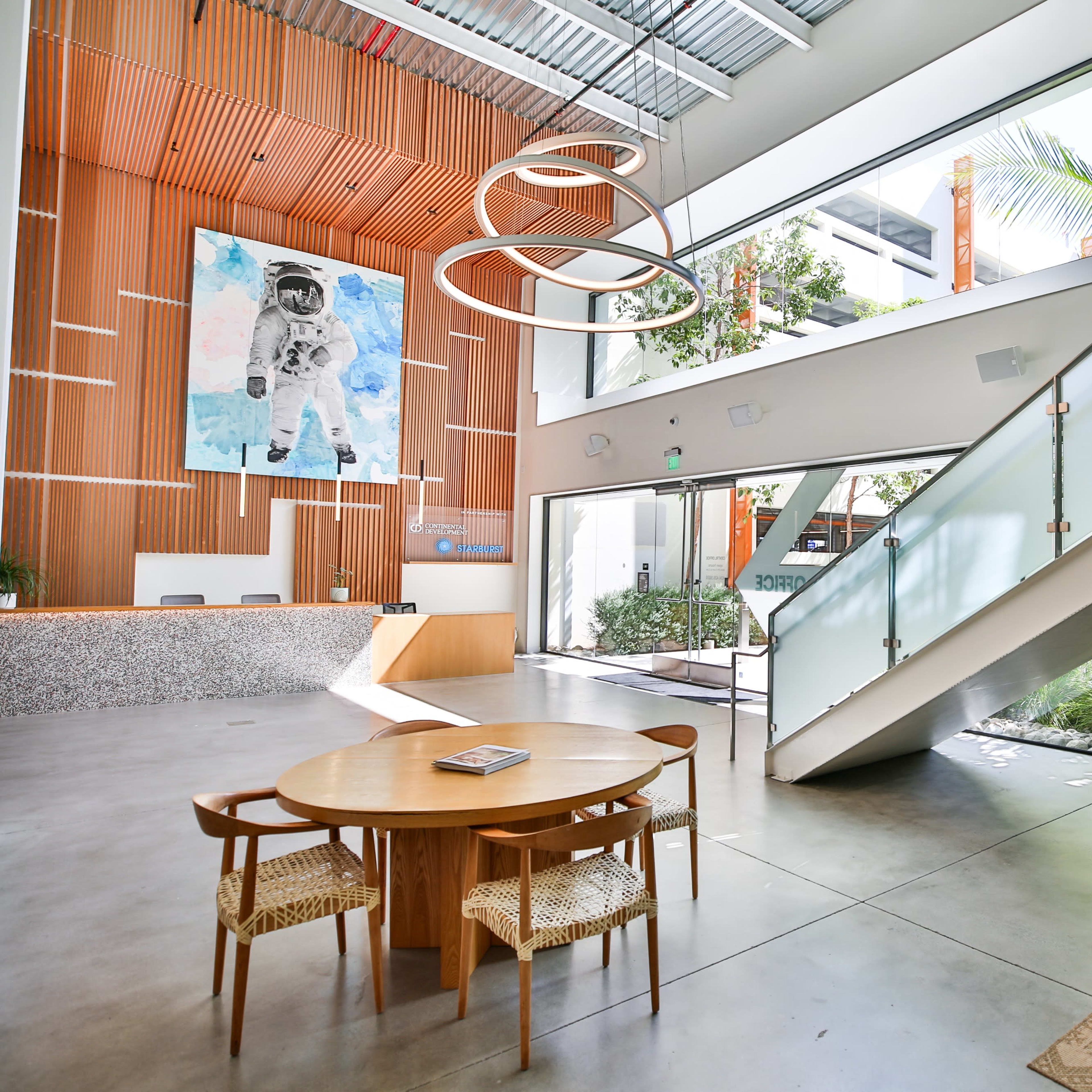 A spacious lobby with a simple circular table, a couch, and a staircase leading to an upper level, characterized by large windows and a prominent artwork of an astronaut on the wall.