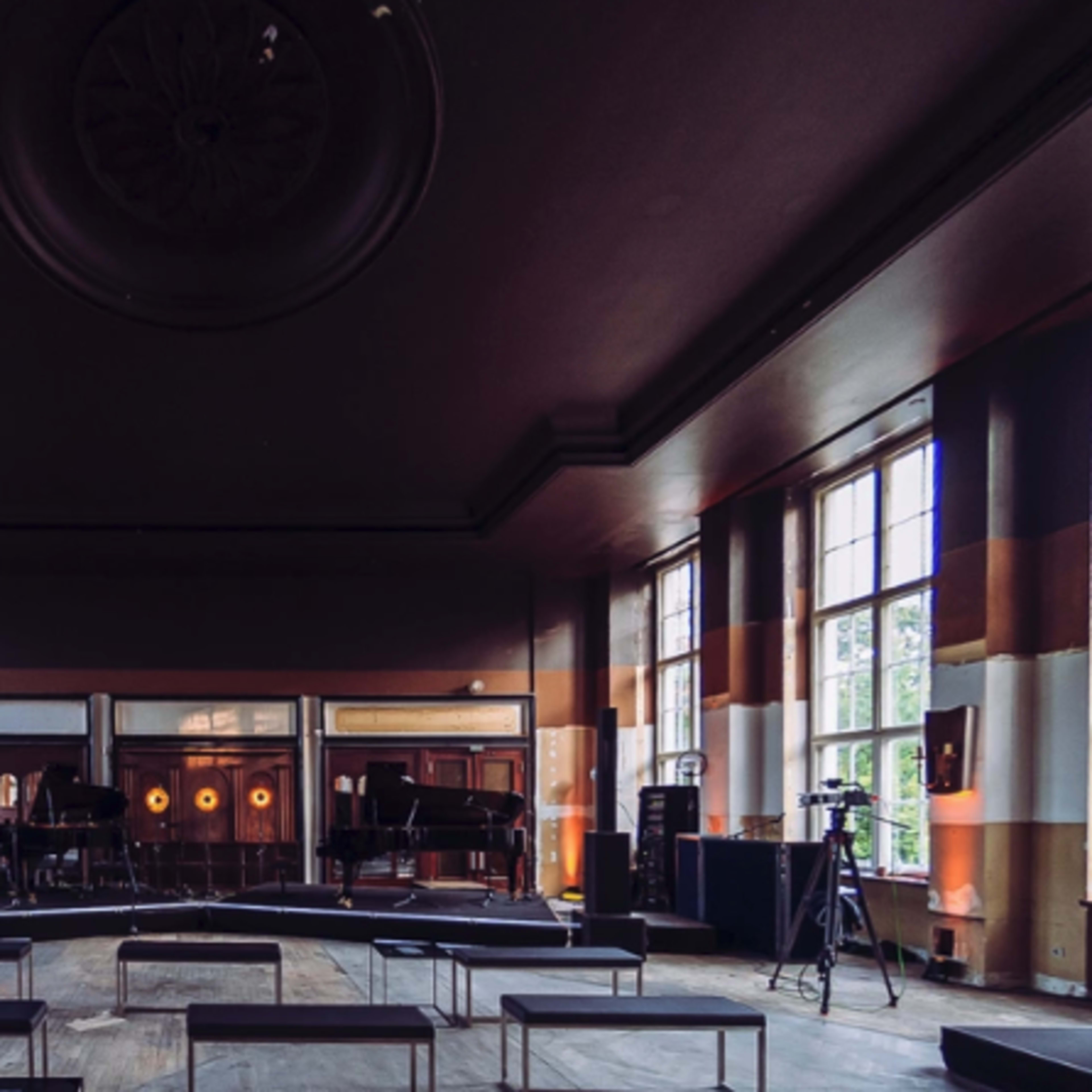 A spacious, well-lit room with large windows, empty seating arrangements, and a small stage set up for a performance.