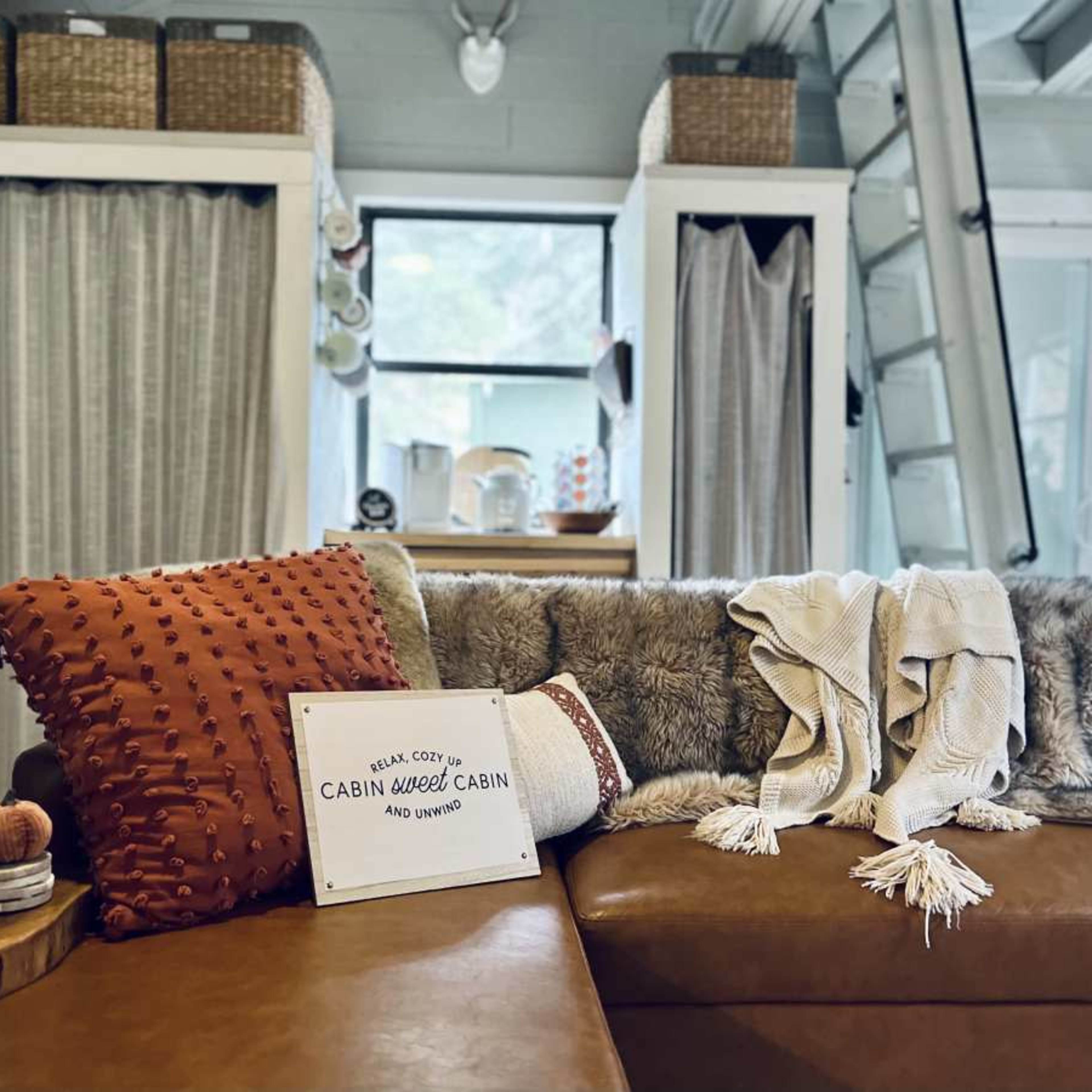 A cozy living room with a leather sofa adorned with decorative pillows and a throw blanket, alongside a sign that reads "CABIN WEED CABIN."