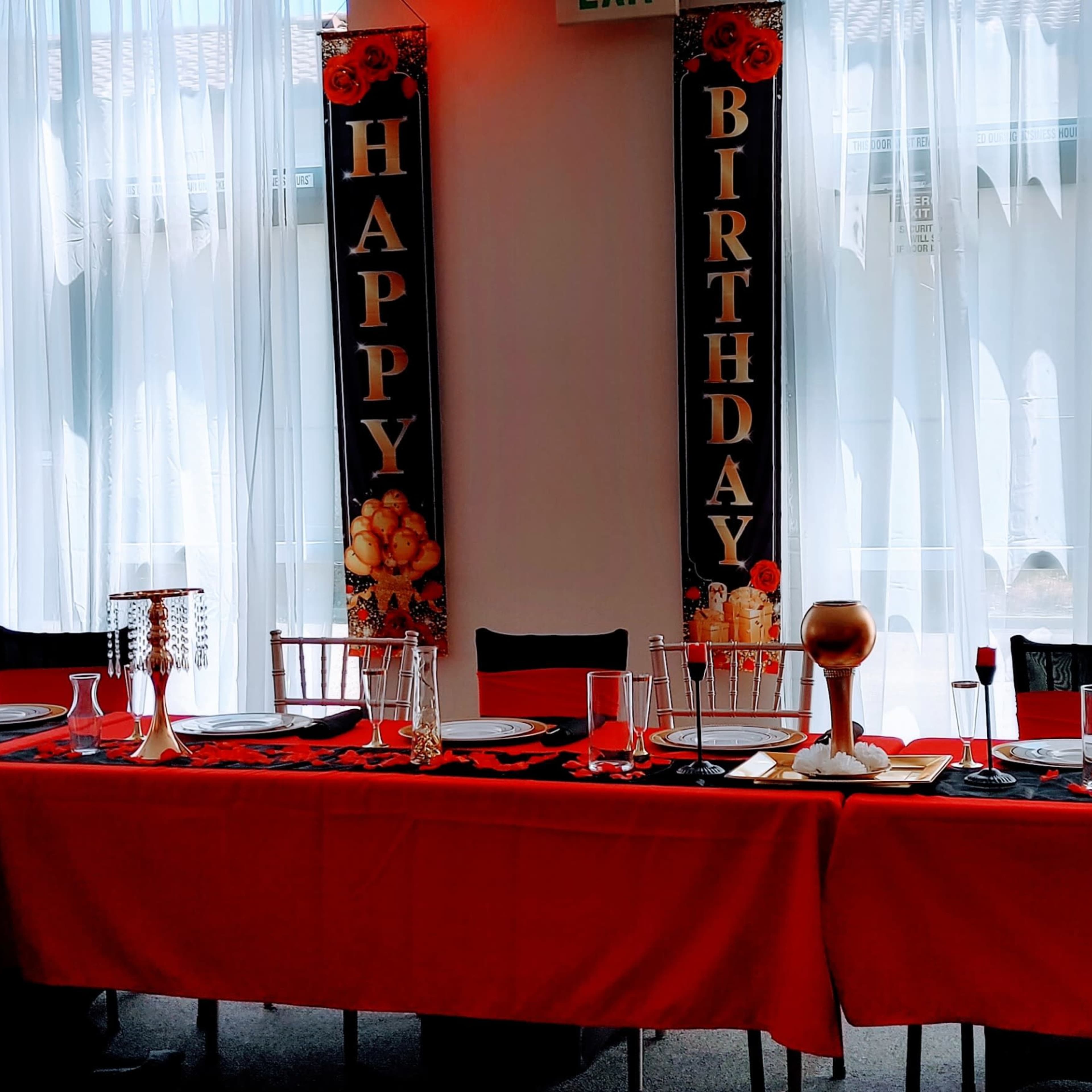 A dining table is set with red tablecloths and black decorations in a room adorned with "HAPPY BIRTHDAY" banners and white curtains.