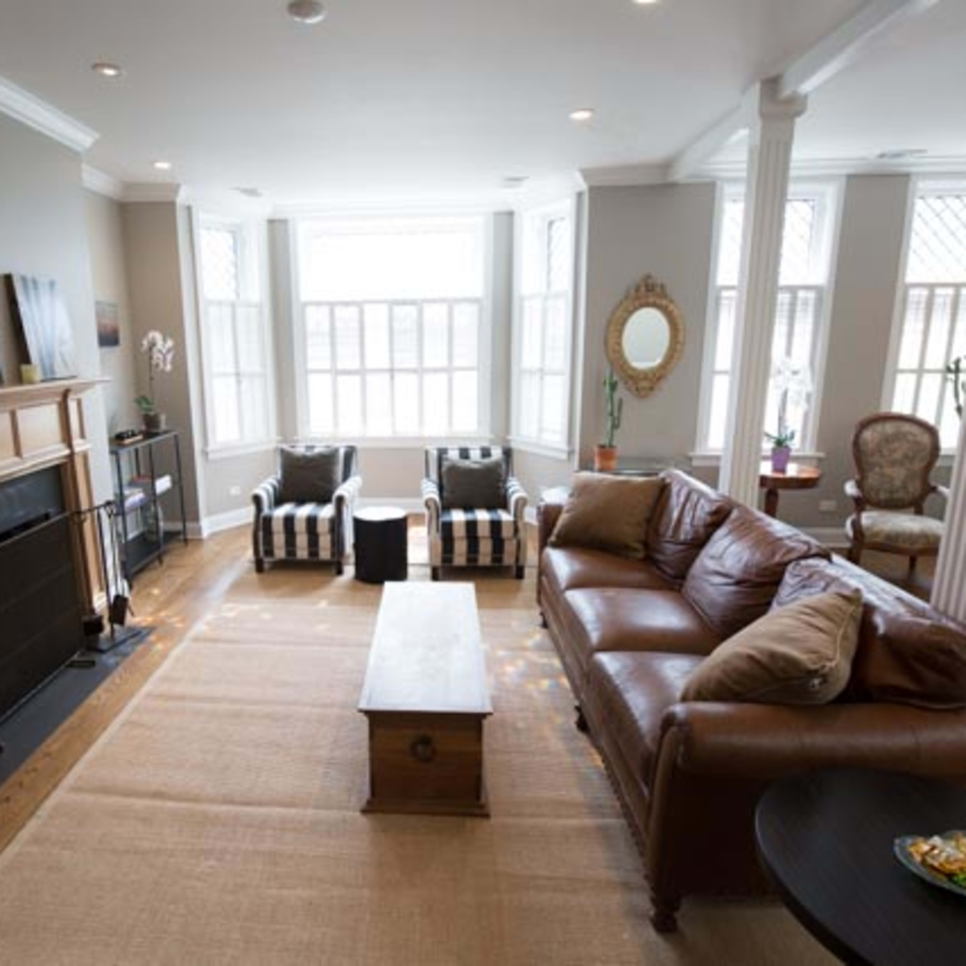 A living room featuring a leather sofa, striped armchairs, a fireplace, and large windows allowing natural light.