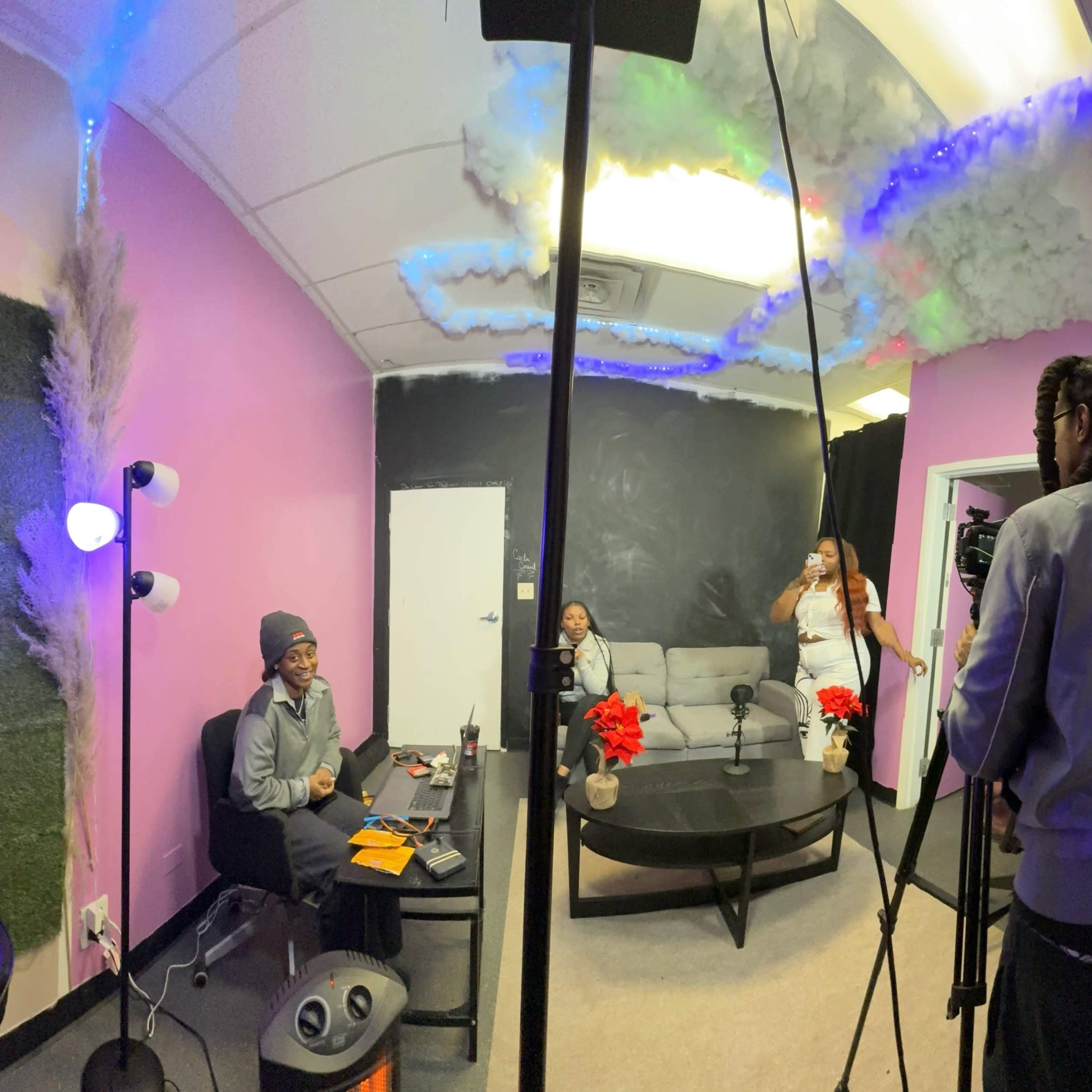 A recording session is taking place in a small studio with pink walls, a black ceiling, and a sofa, where two people are seated at a table while another person stands nearby holding a camera.