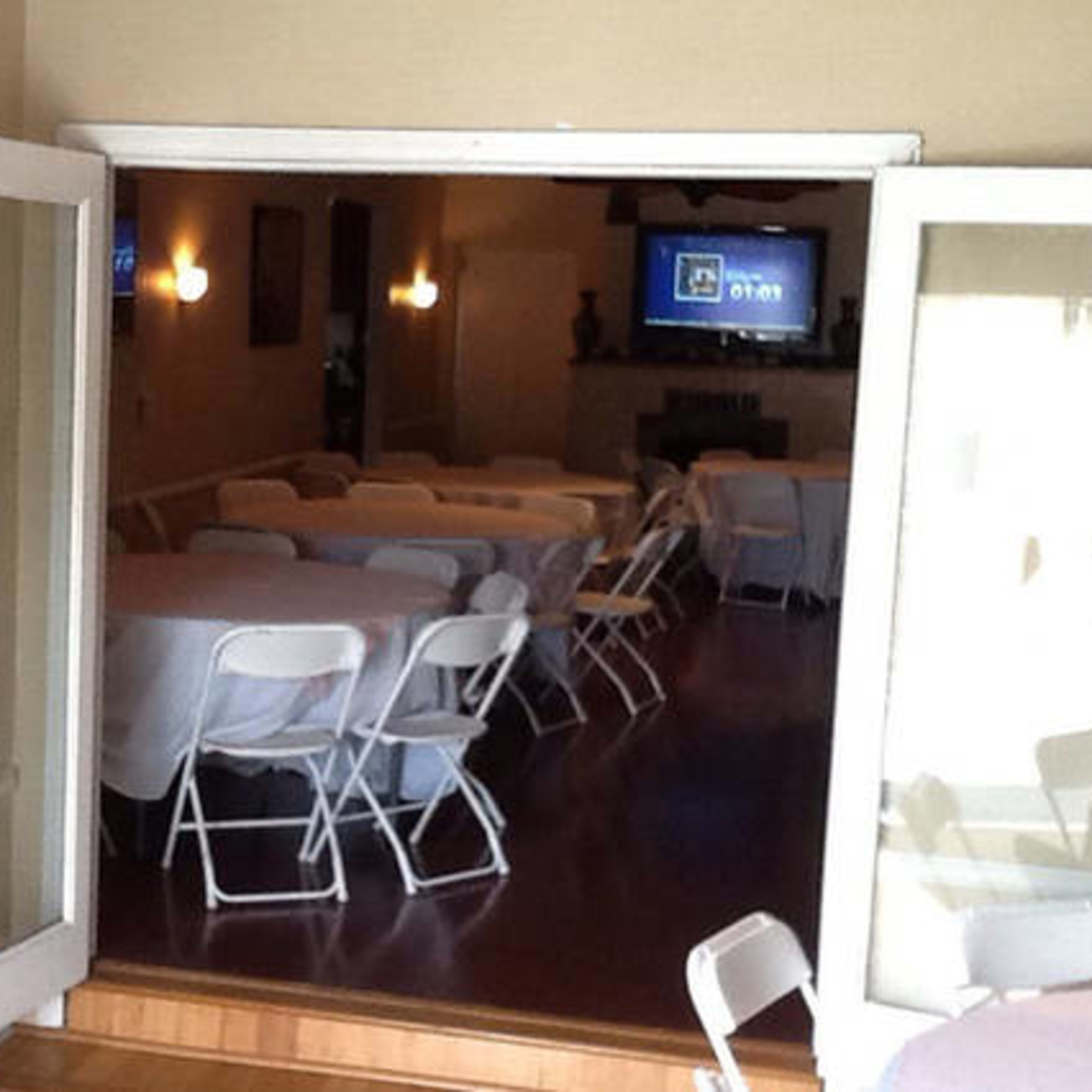 An interior room with several tables covered in white tablecloths and arranged with plastic chairs, visible through open double doors.