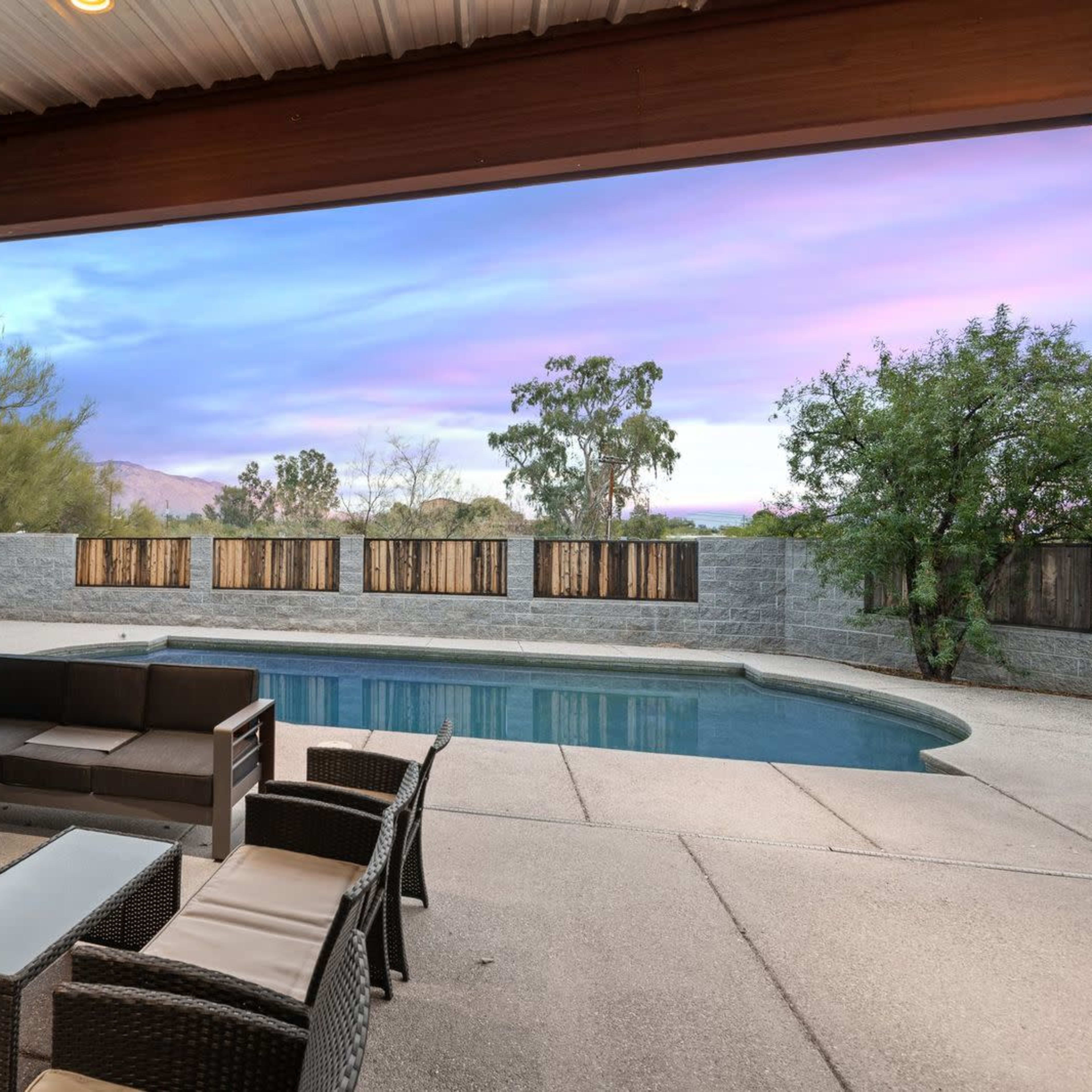 A backyard with a swimming pool, outdoor seating, and a fence surrounded by greenery under a colorful sunset sky.