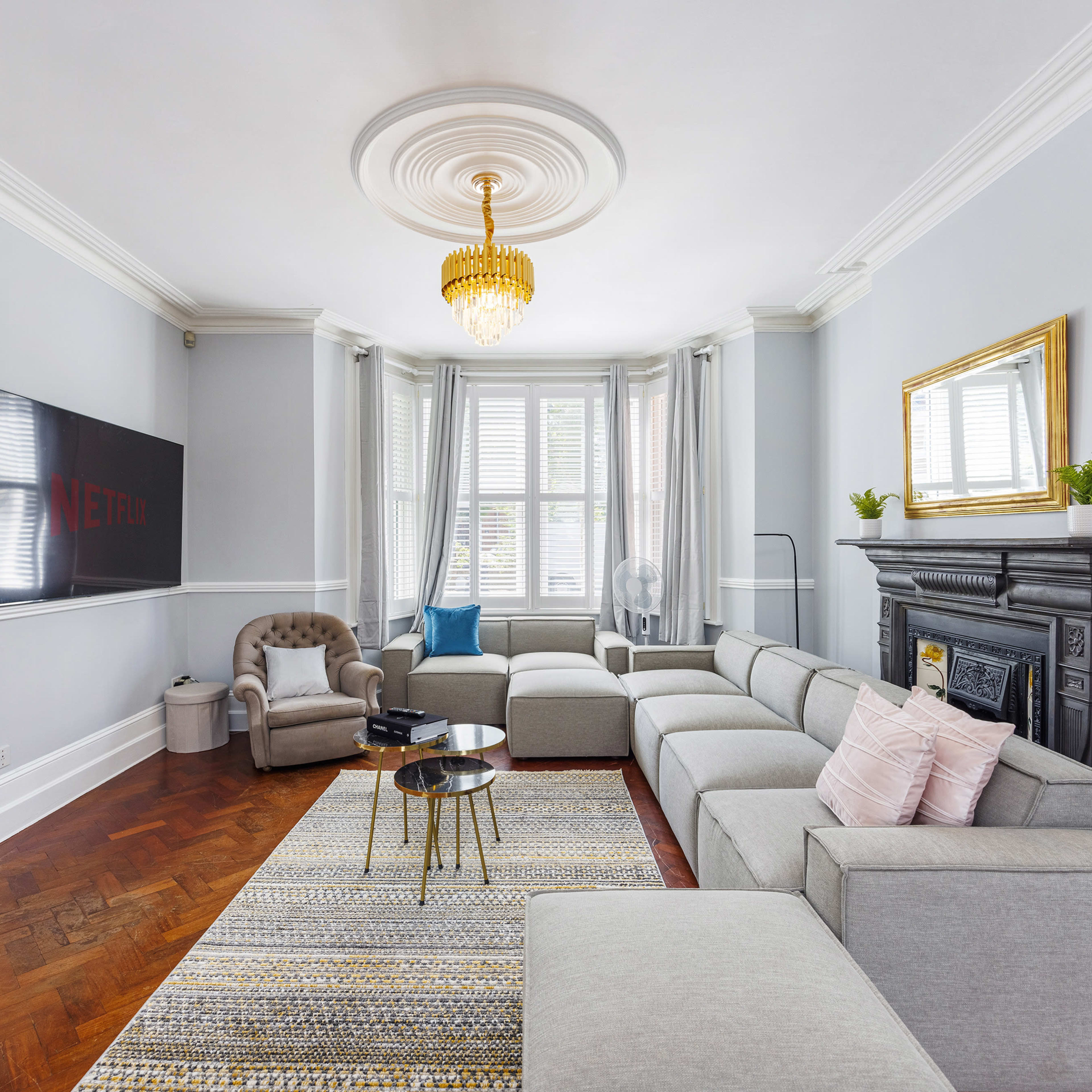The living room features a modern sectional sofa, a round coffee table, and a wall-mounted TV, all set against a backdrop of elegant moldings and a decorative fireplace.