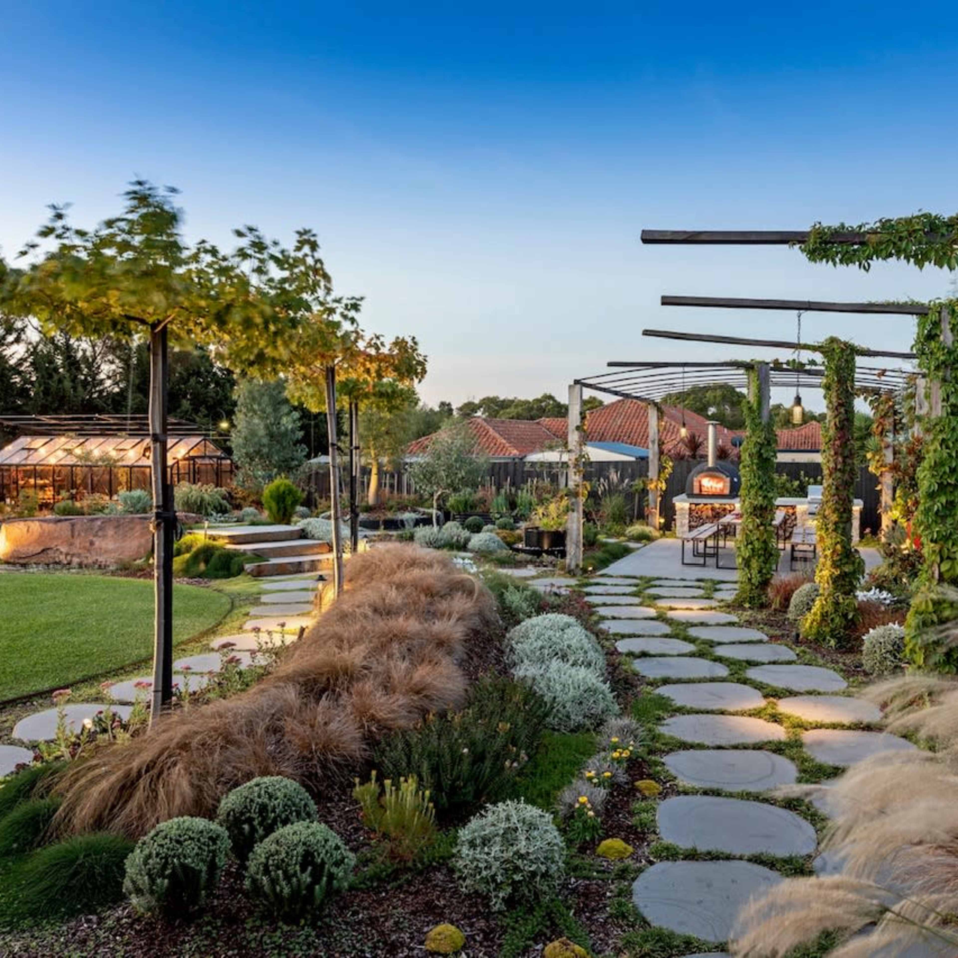 A landscaped garden features stone pathways, greenery, and a covered seating area under a blue sky.