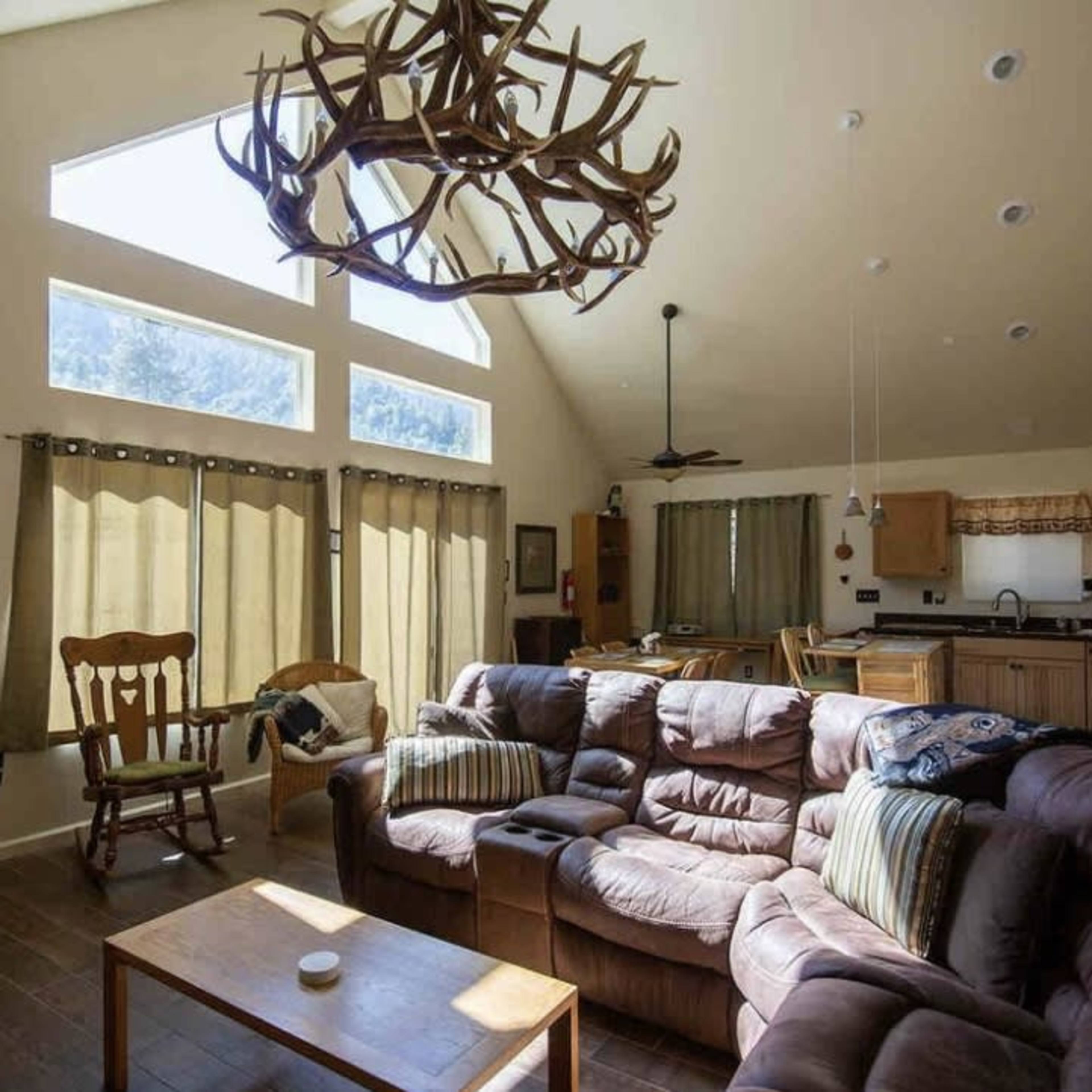 A spacious living room with large windows, a brown sectional sofa, and a decorative antler chandelier.