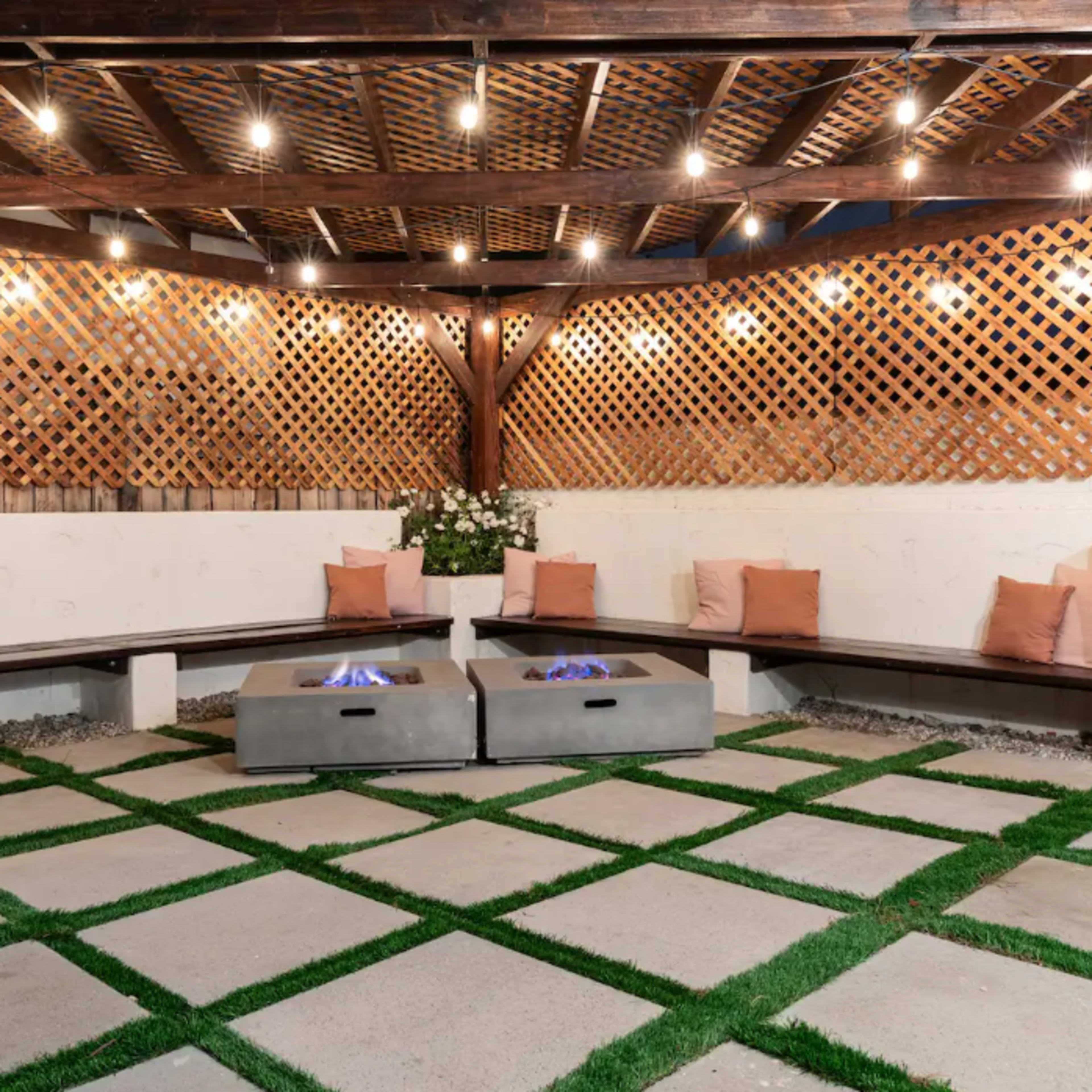 An outdoor seating area with a firepit surrounded by benches, illuminated by string lights overhead and featuring a lattice backdrop.