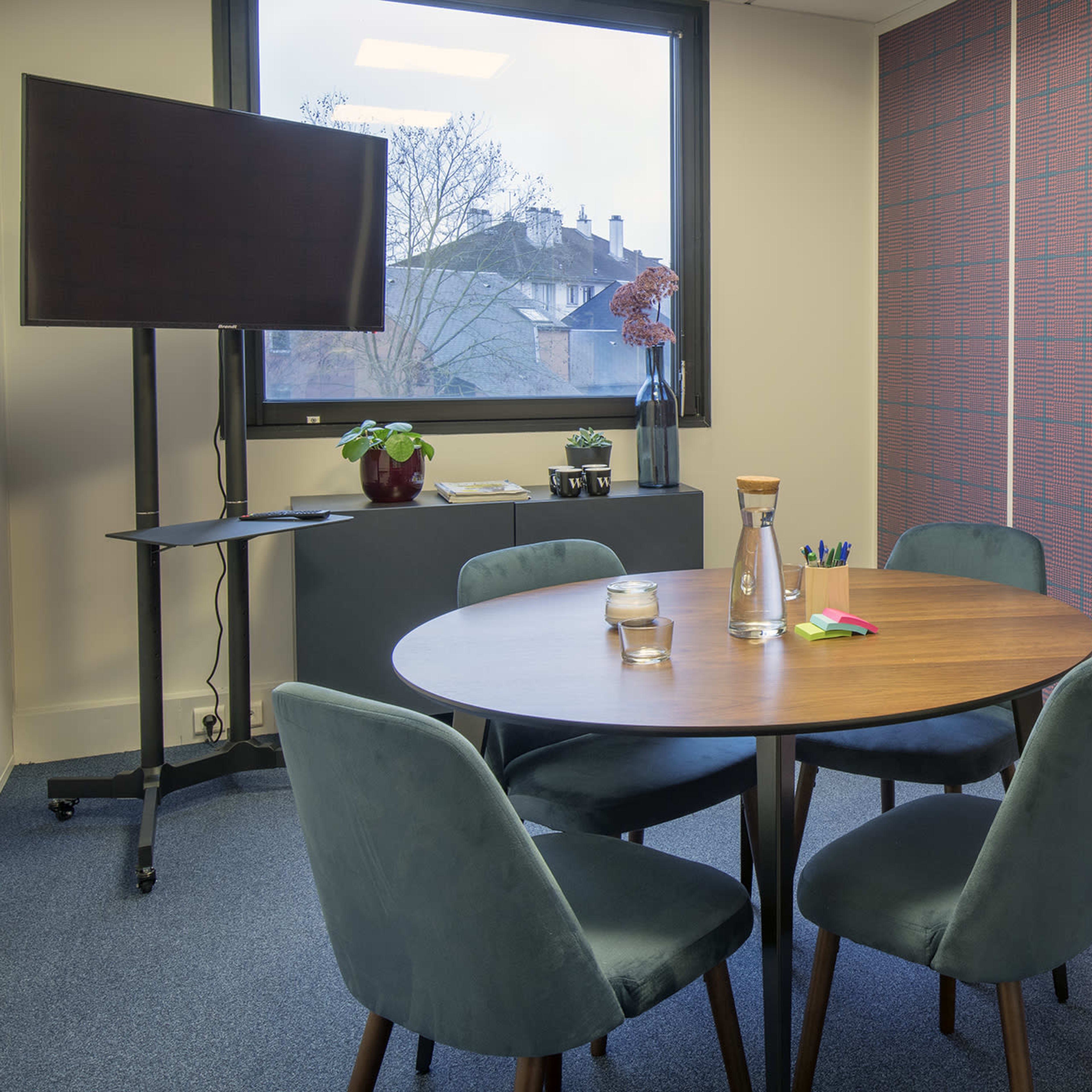 A round wooden table is surrounded by four green upholstered chairs in a small meeting room with a television on a stand and a patterned wall.