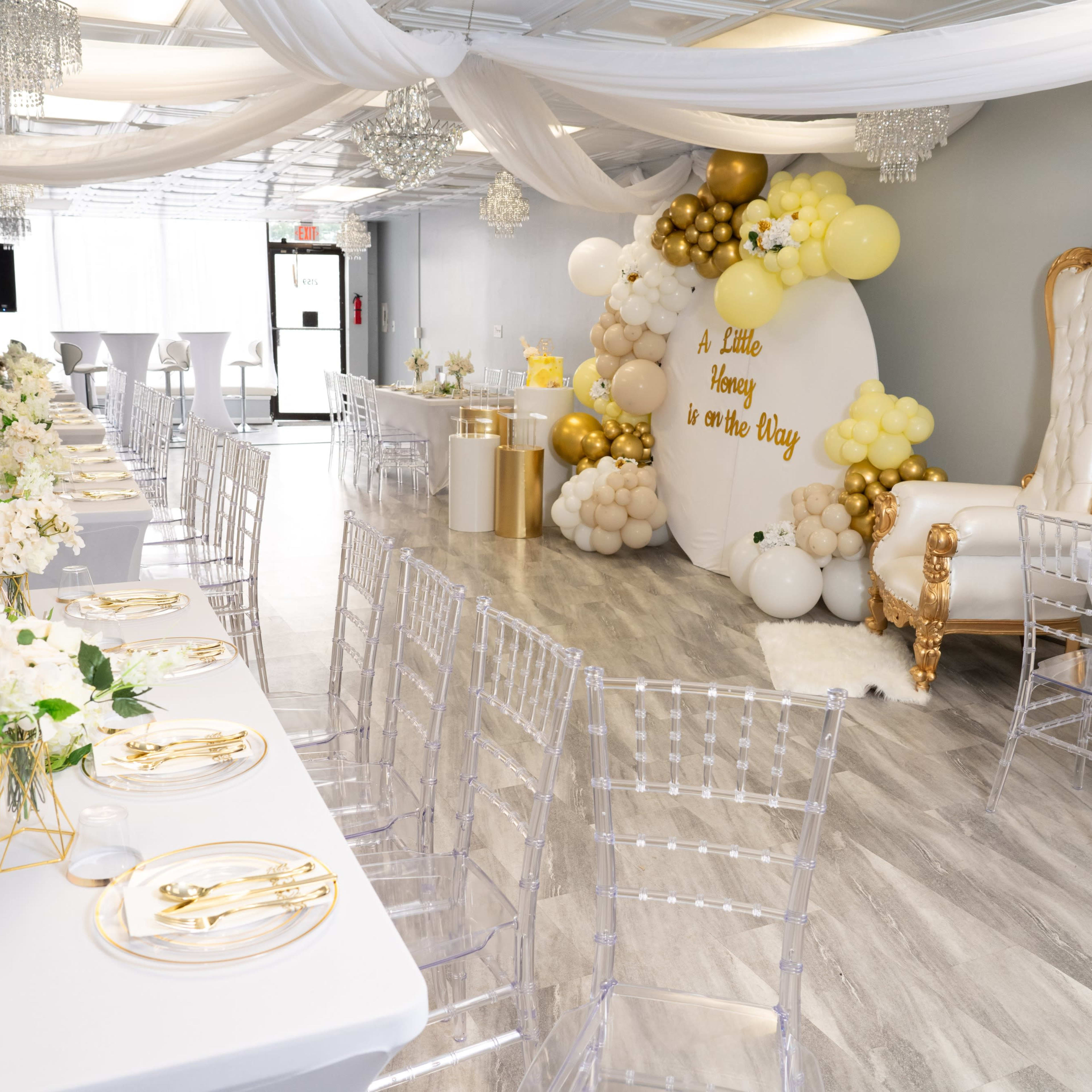A decorated event space featuring tables with white tablecloths and gold accents, a themed balloon backdrop, and a large ornate chair.