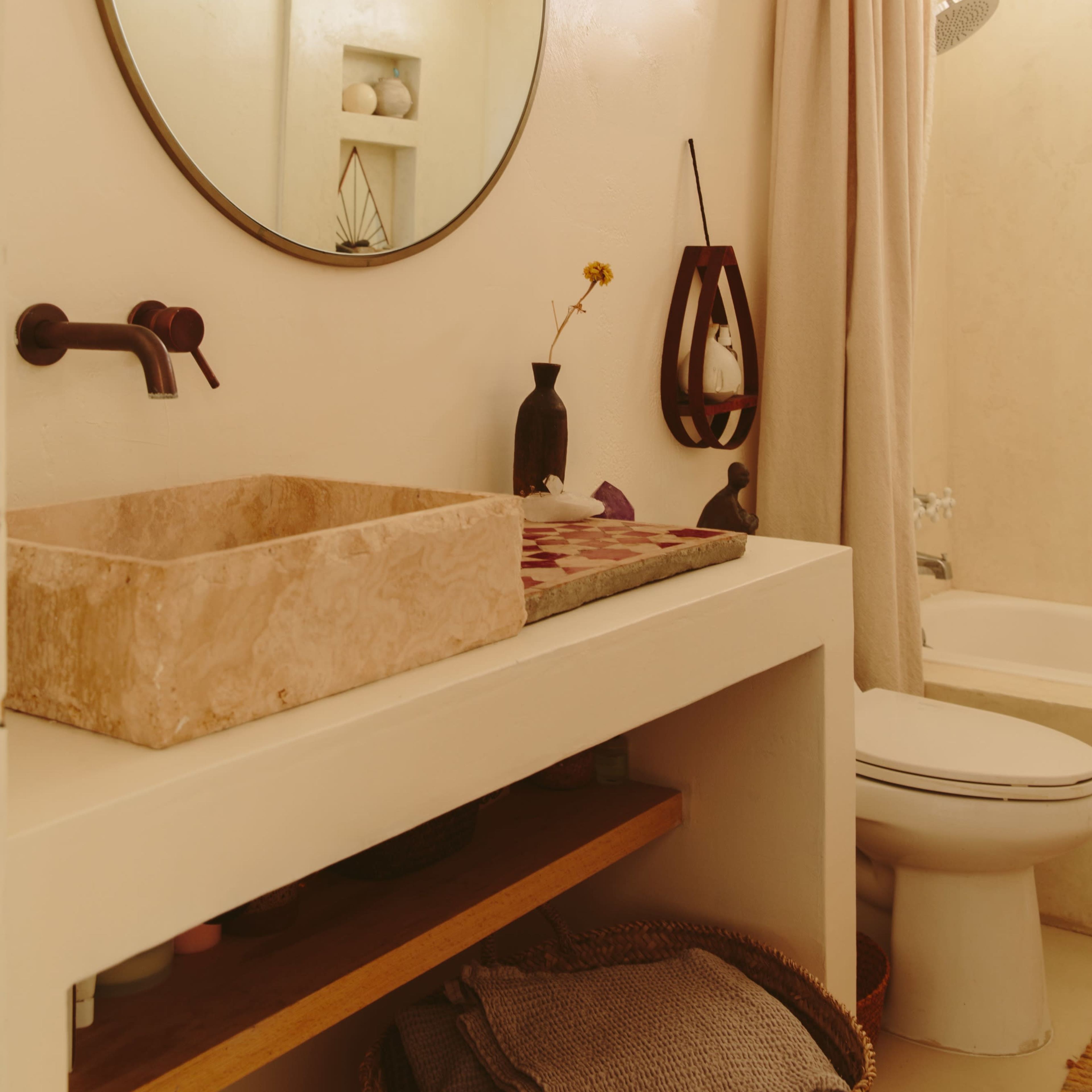 A modern bathroom features a circular mirror above a stone sink, with a wooden shelf and a bathtub visible in the background.