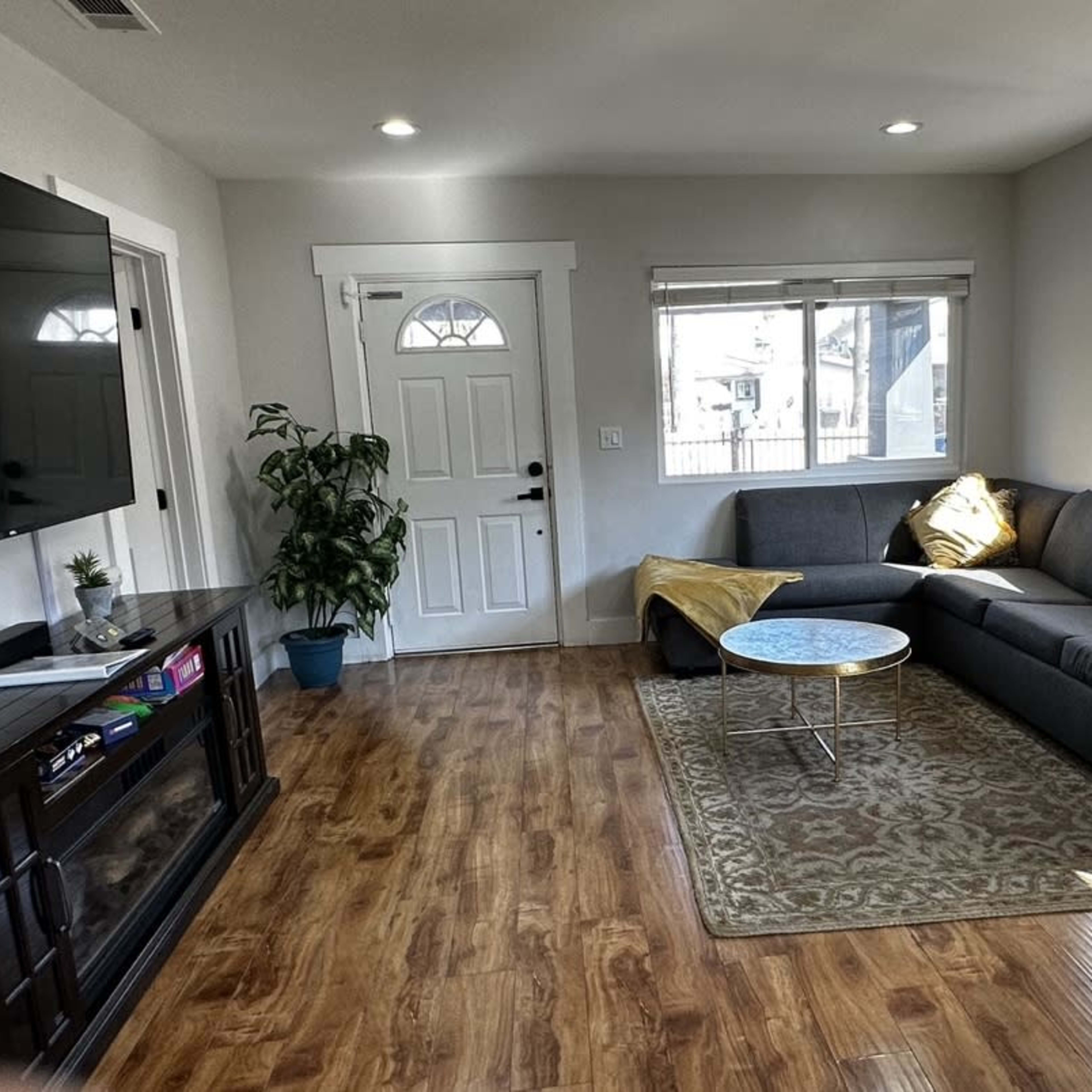 The room features a gray sectional sofa, a small coffee table, a television mounted on the wall, and a front door leading outside.