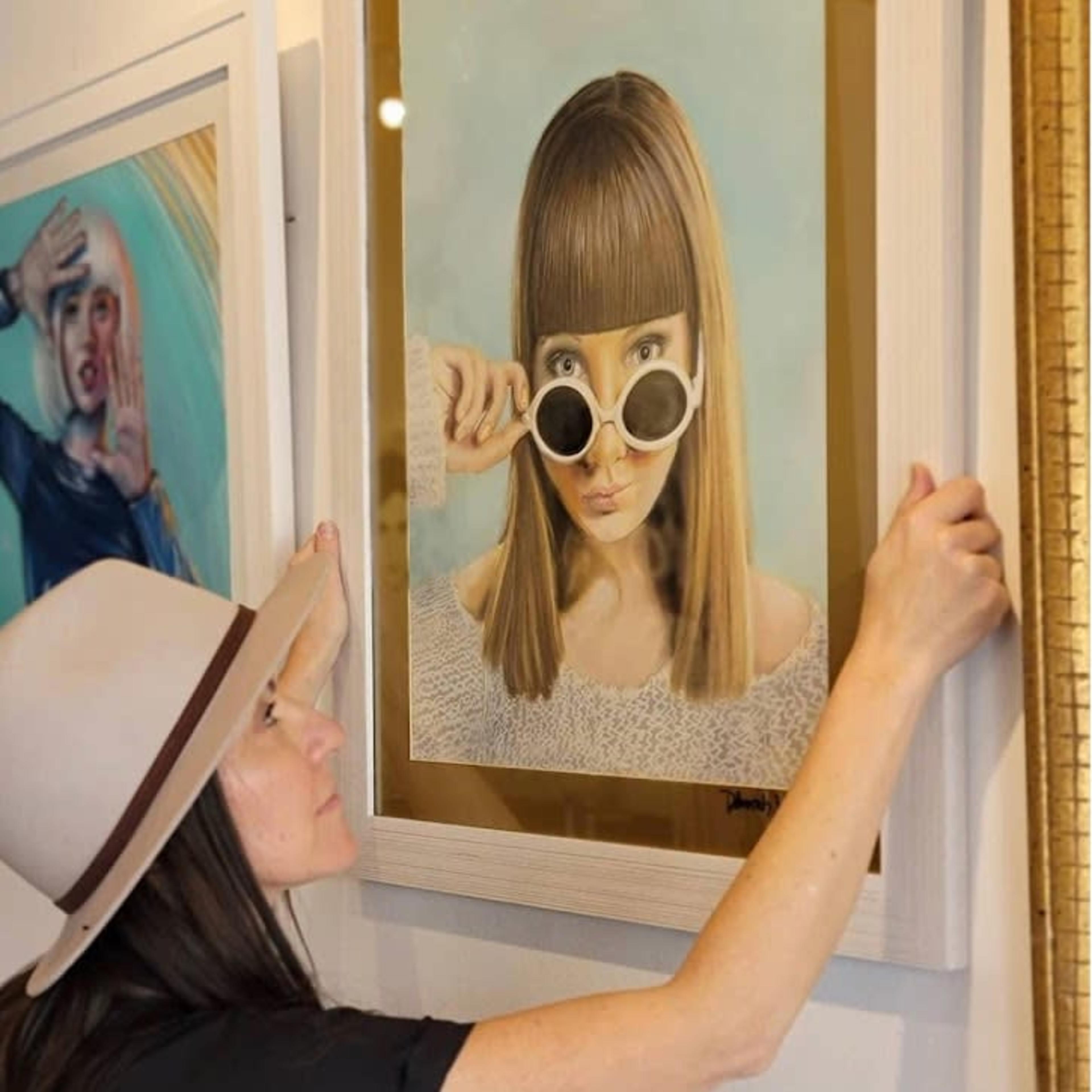 A woman in a hat adjusts a framed portrait of a model wearing sunglasses on the wall of an art gallery.