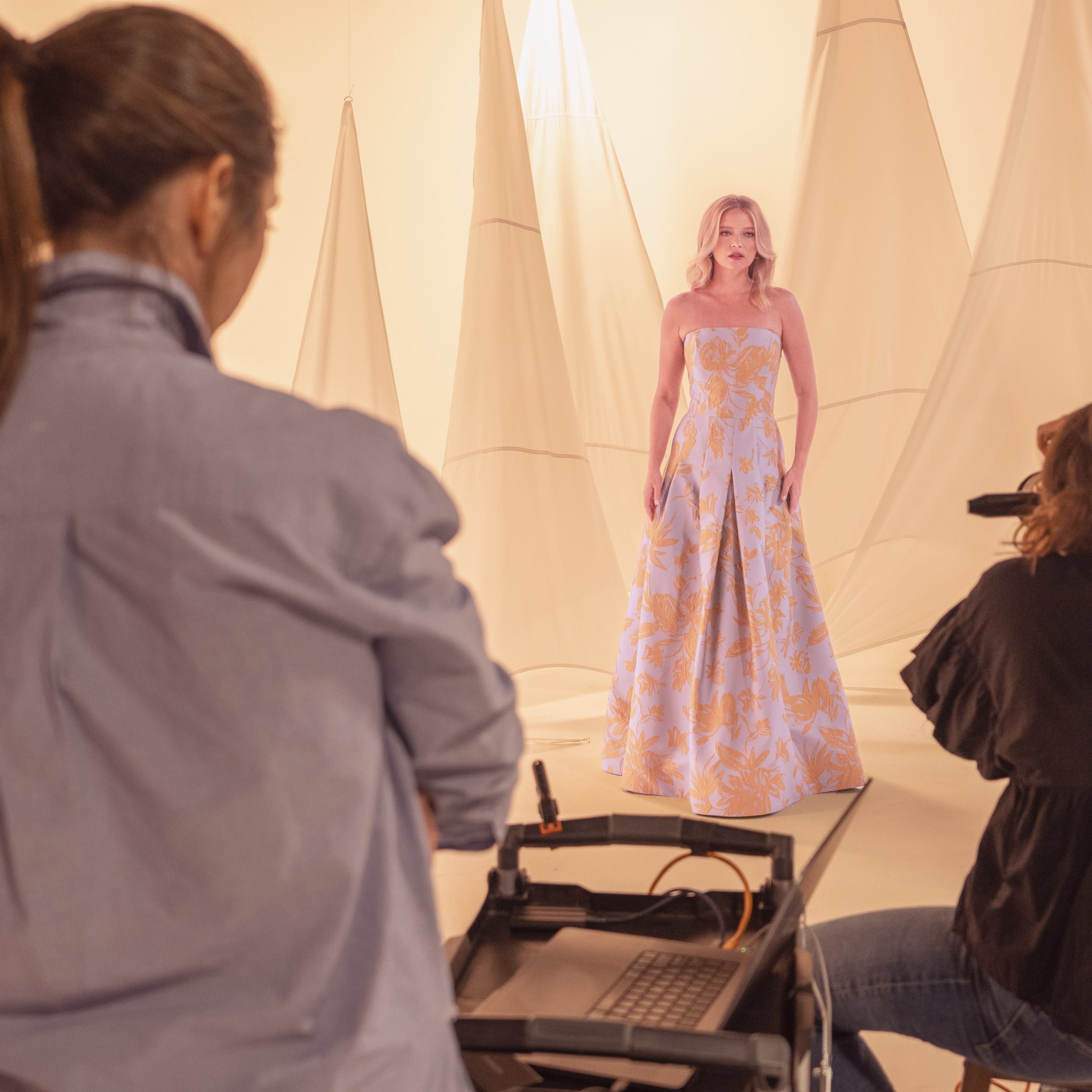A model in a floral dress poses for a photoshoot in a studio with fabric backdrops.