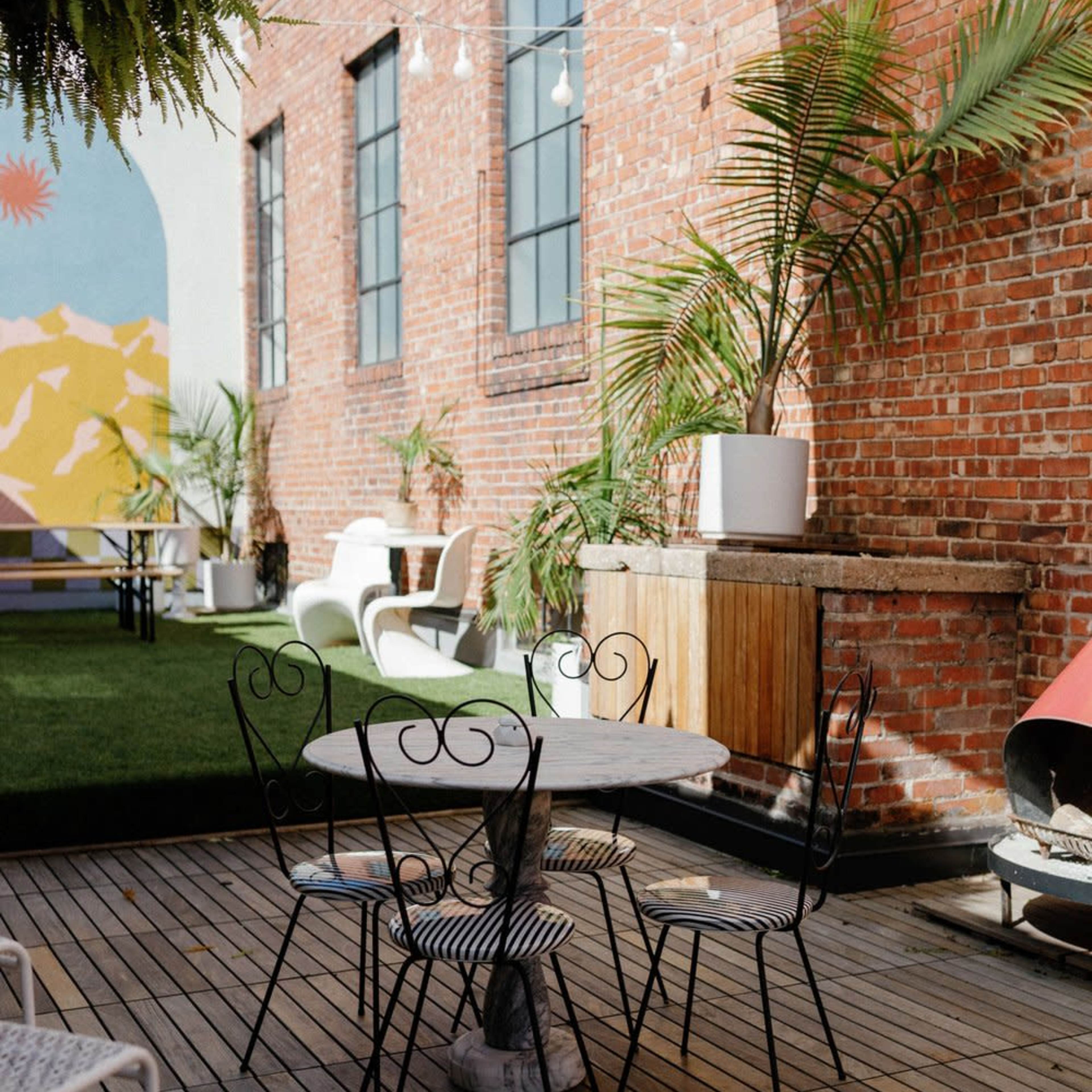 A small outdoor seating area features a round table with four wrought iron chairs on a wooden deck, surrounded by potted plants and a brick wall.